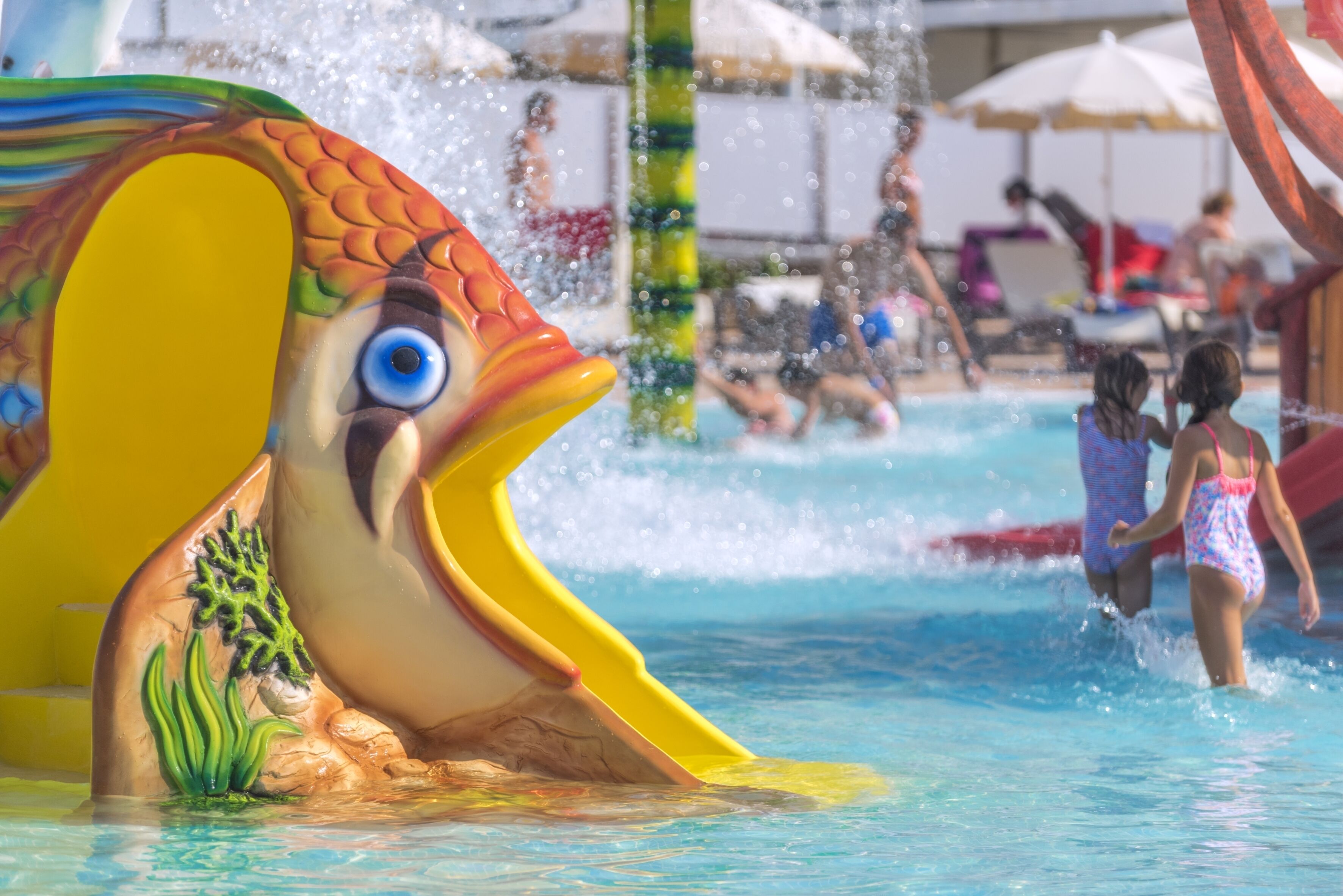 A water slide shaped like a fish with a large blue eye, surrounded by a pool area with people and umbrellas.