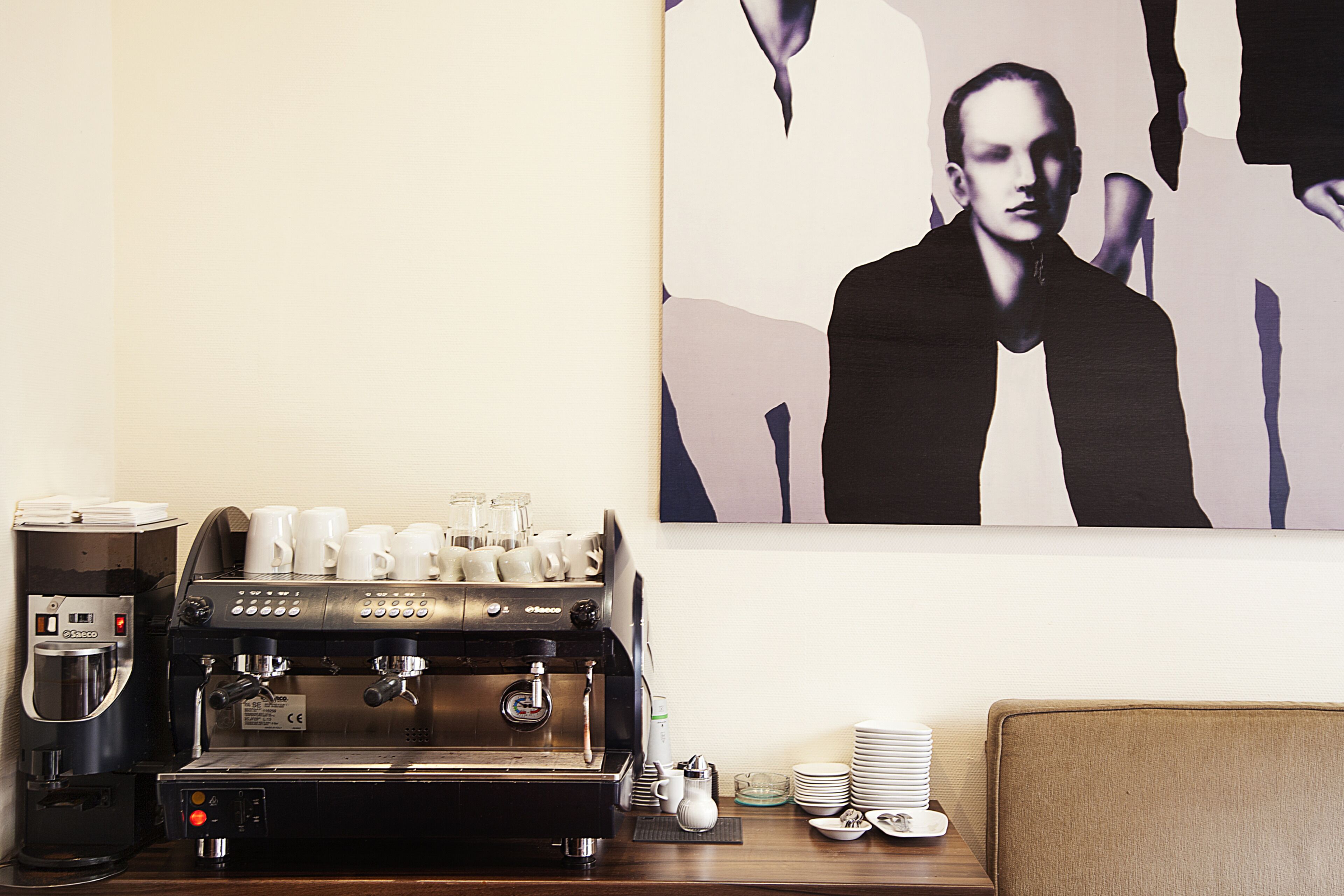 A coffee machine with multiple cups and a coffee dispenser, a stack of plates, and a framed artwork on the wall.