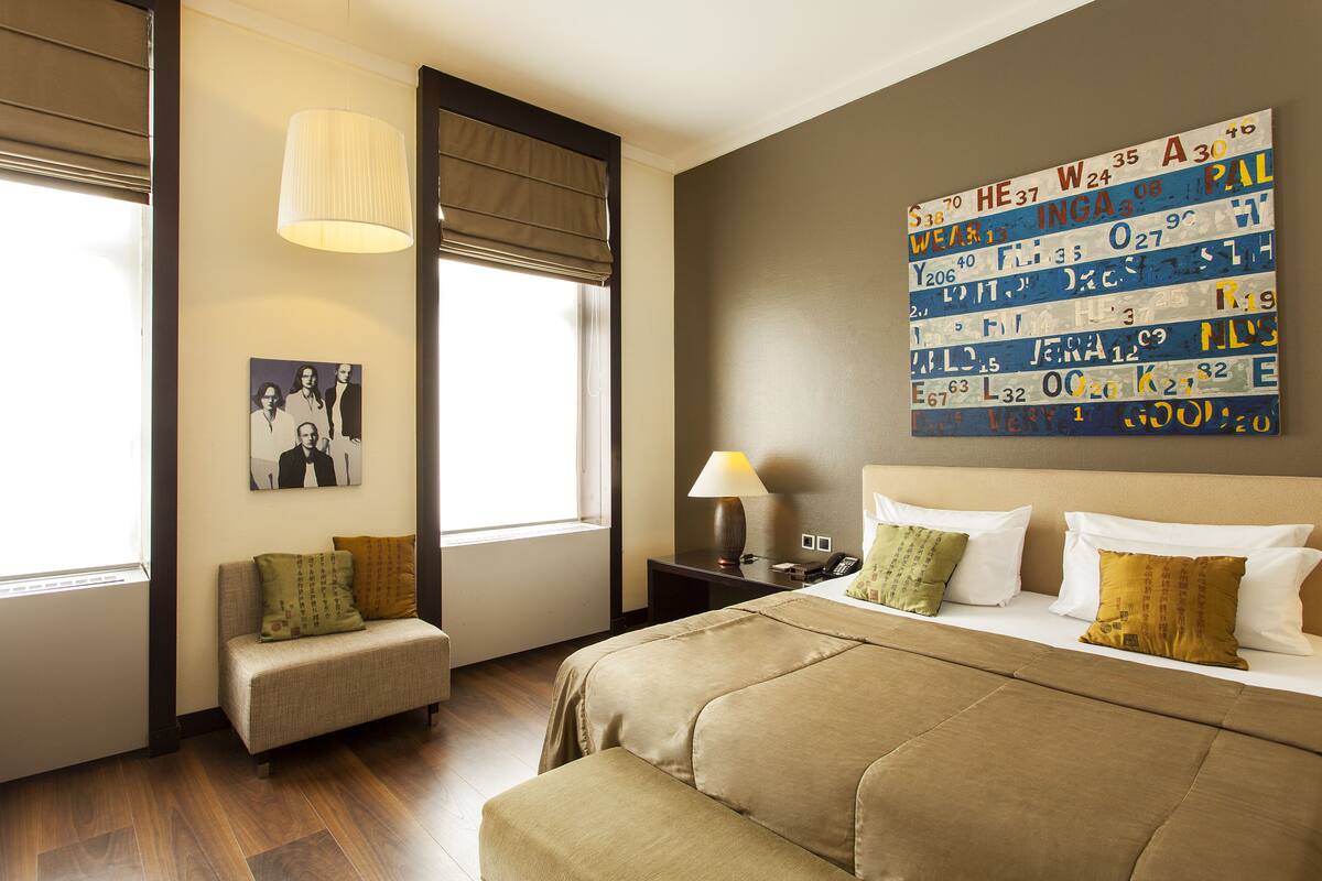 Ein Hotelzimmer mit Bett, Sofa, Lampe, Gemälde und einem Fenster mit Jalousien.