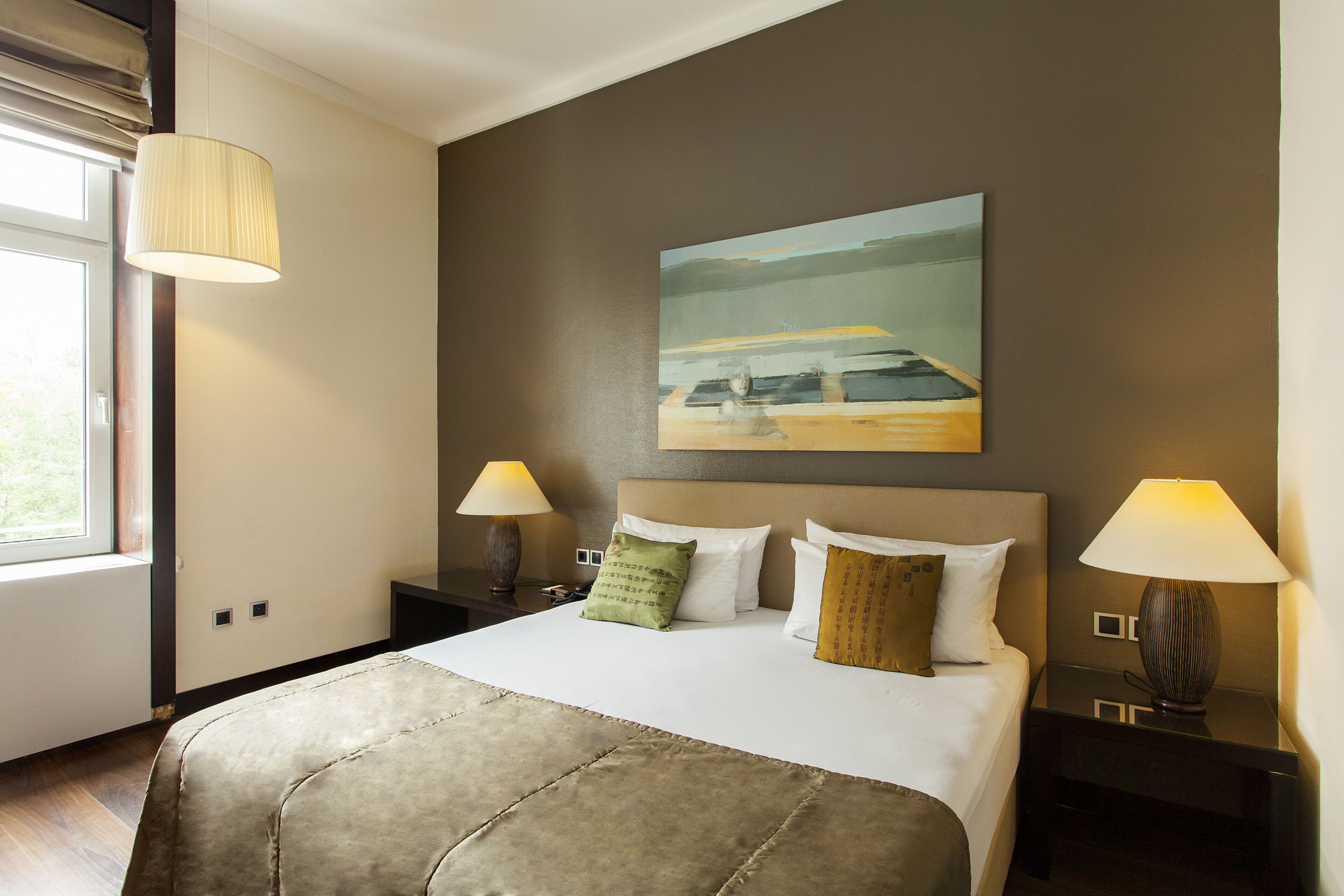 Una habitación de hotel con una cama grande, dos mesitas de noche con lámparas, un cuadro en la pared y una ventana con cortinas.