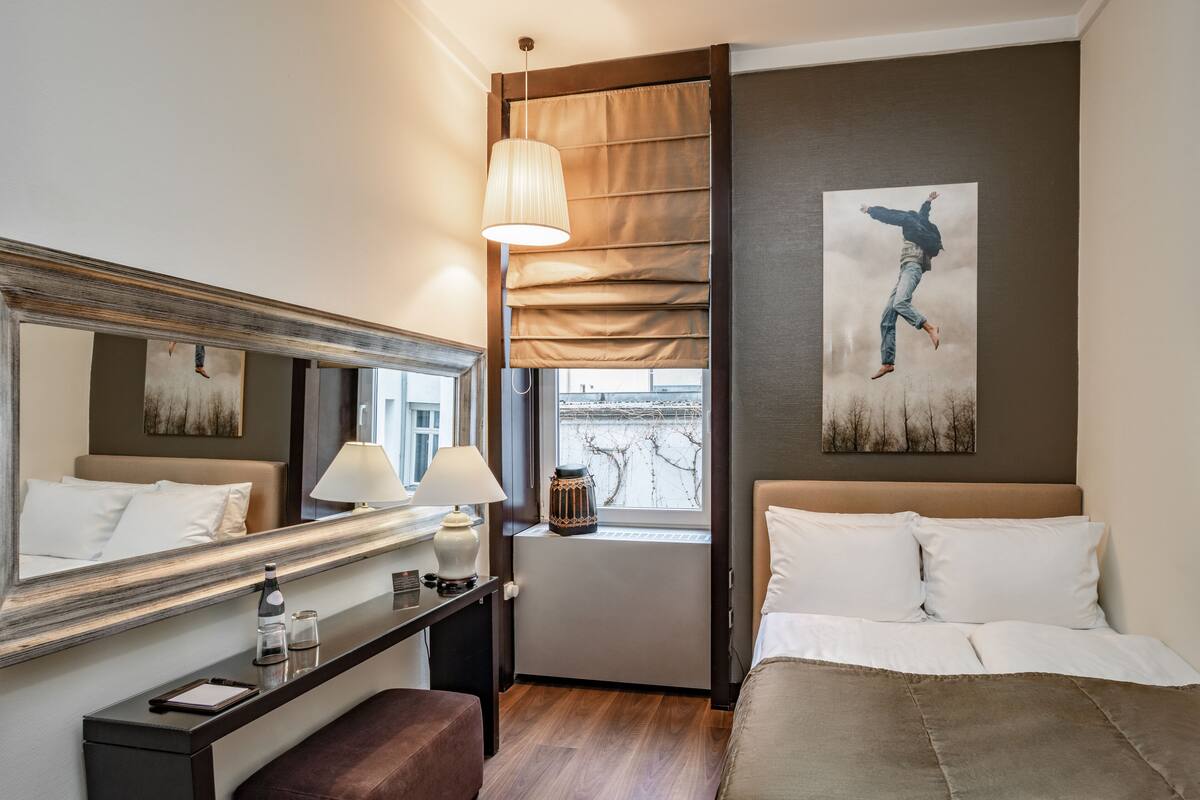 Ein Hotelzimmer mit einem großen Bett, einem Nachttisch mit Lampe, einem Fenster mit Jalousien, einem Heizkörper und einem Bild an der Wand.