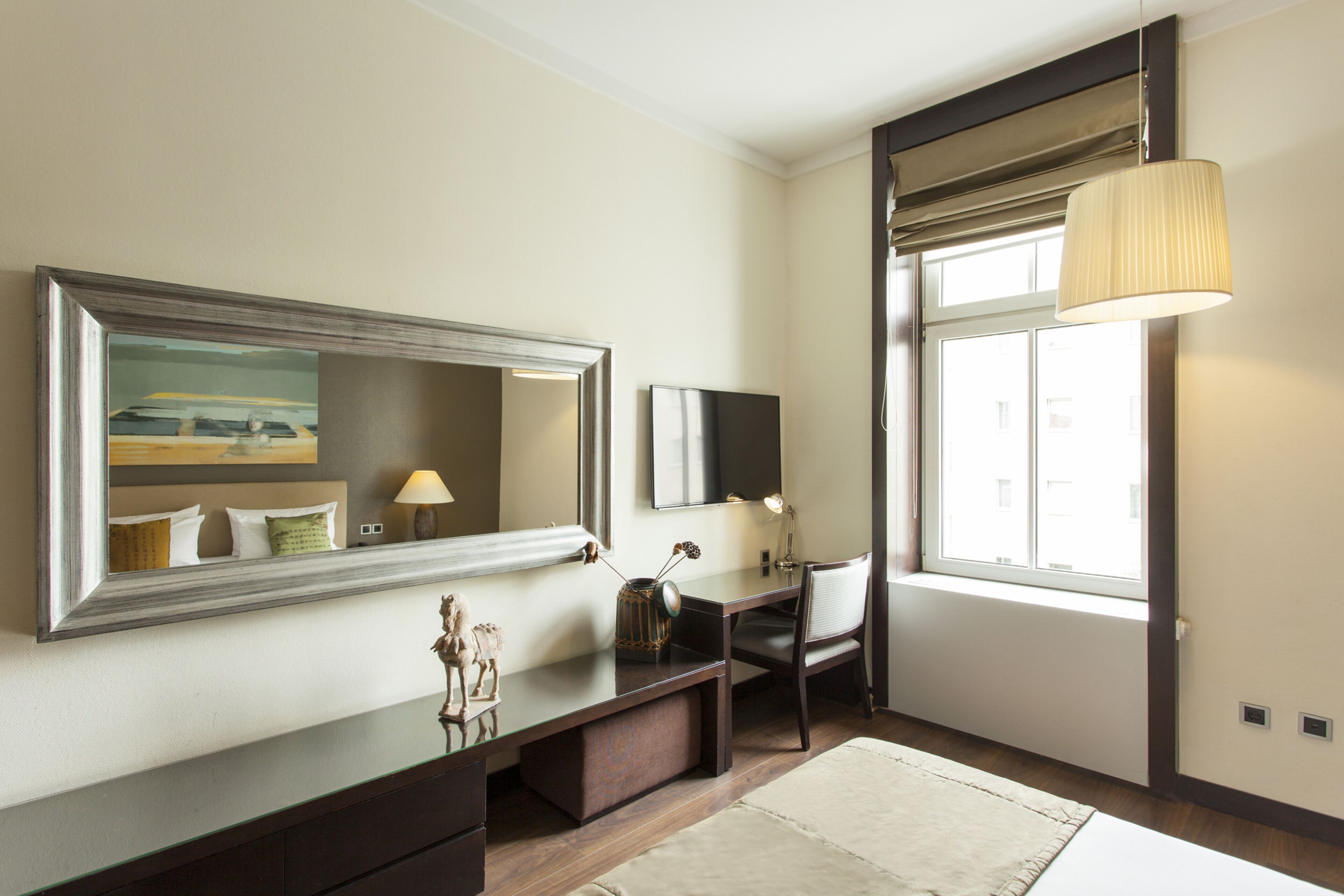 Ein Hotelzimmer mit einem großen Spiegel, einem Schreibtisch mit Stuhl, einer Lampe und einem Fenster mit Blick.
