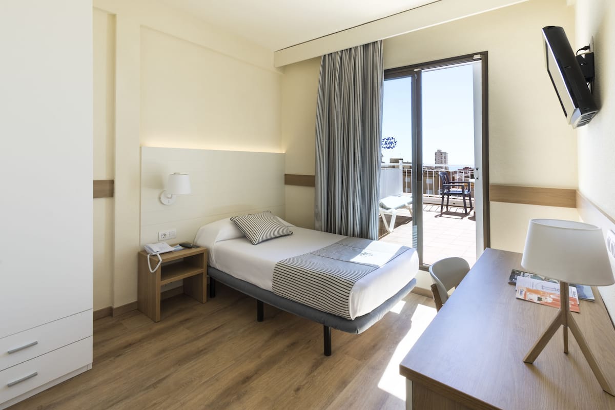 A hotel room with a bed, bedside table, desk, chair, and a view of the city through a large window.