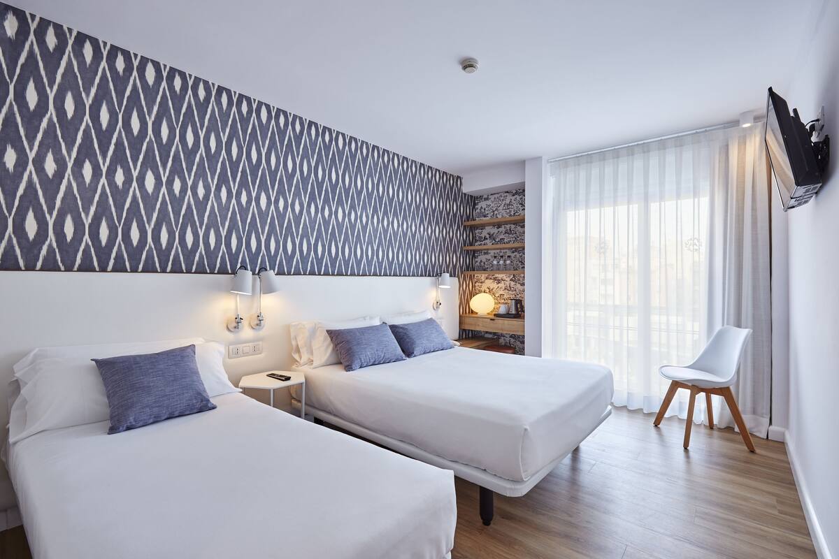 A hotel room with two beds, a large patterned wall, a TV mounted on the wall, and a window with sheer curtains.