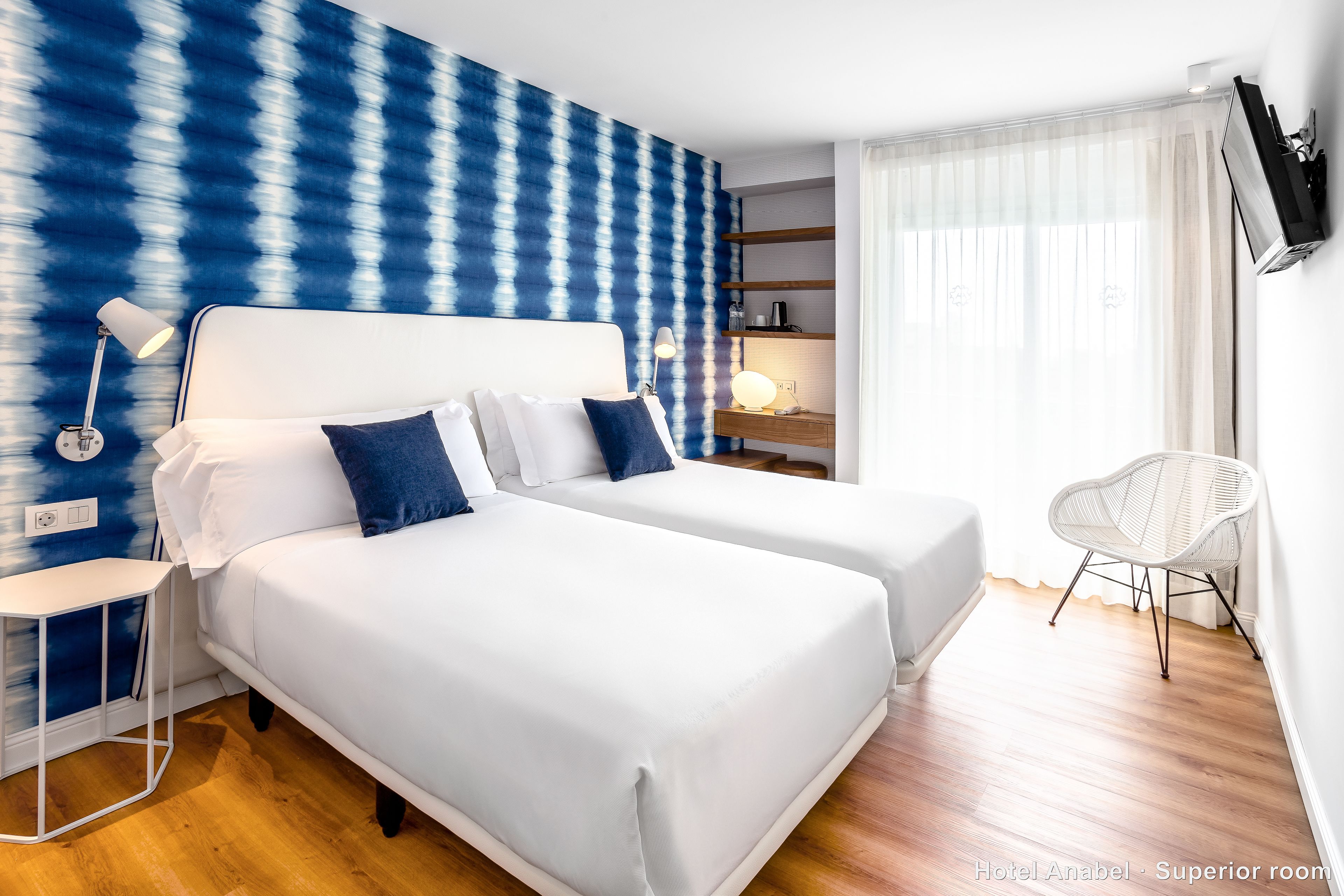 A hotel room with two beds, a white lamp, a small table, a chair, and a TV mounted on the wall.