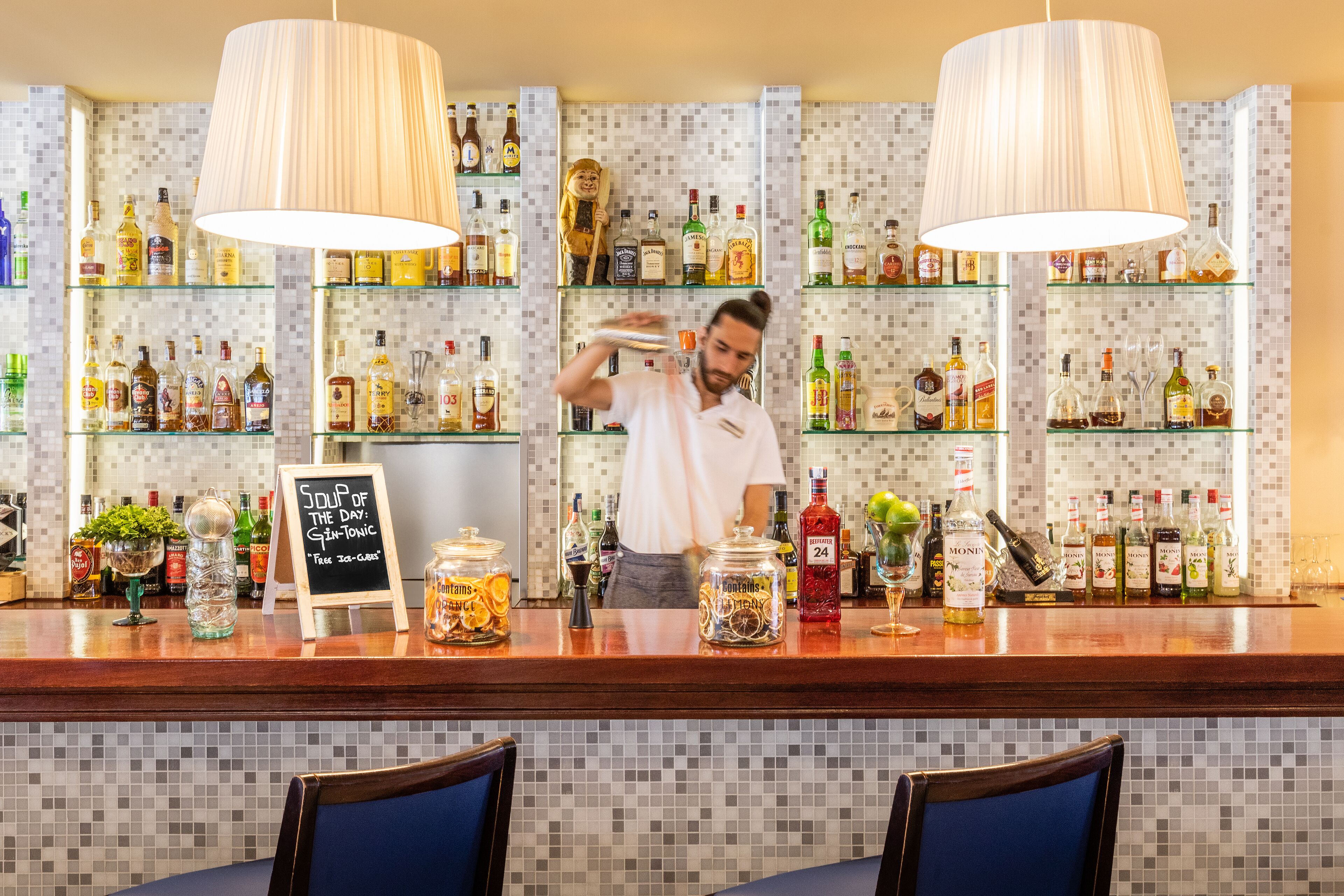 Zona de bar con estantes llenos de botellas de licores, un menú en la pizarra y una persona preparando bebidas.