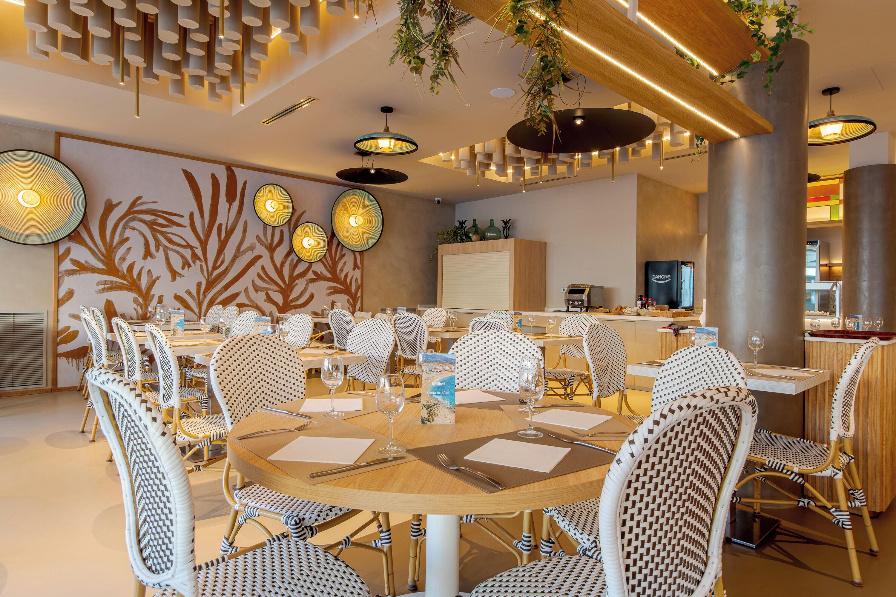 A modern dining area with round tables and wicker chairs, a kitchen in the background, and decorative wall art.