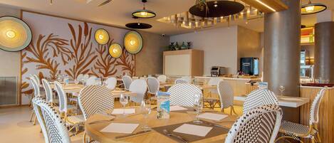 A modern dining area with round tables and wicker chairs, a kitchen in the background, and decorative wall art.