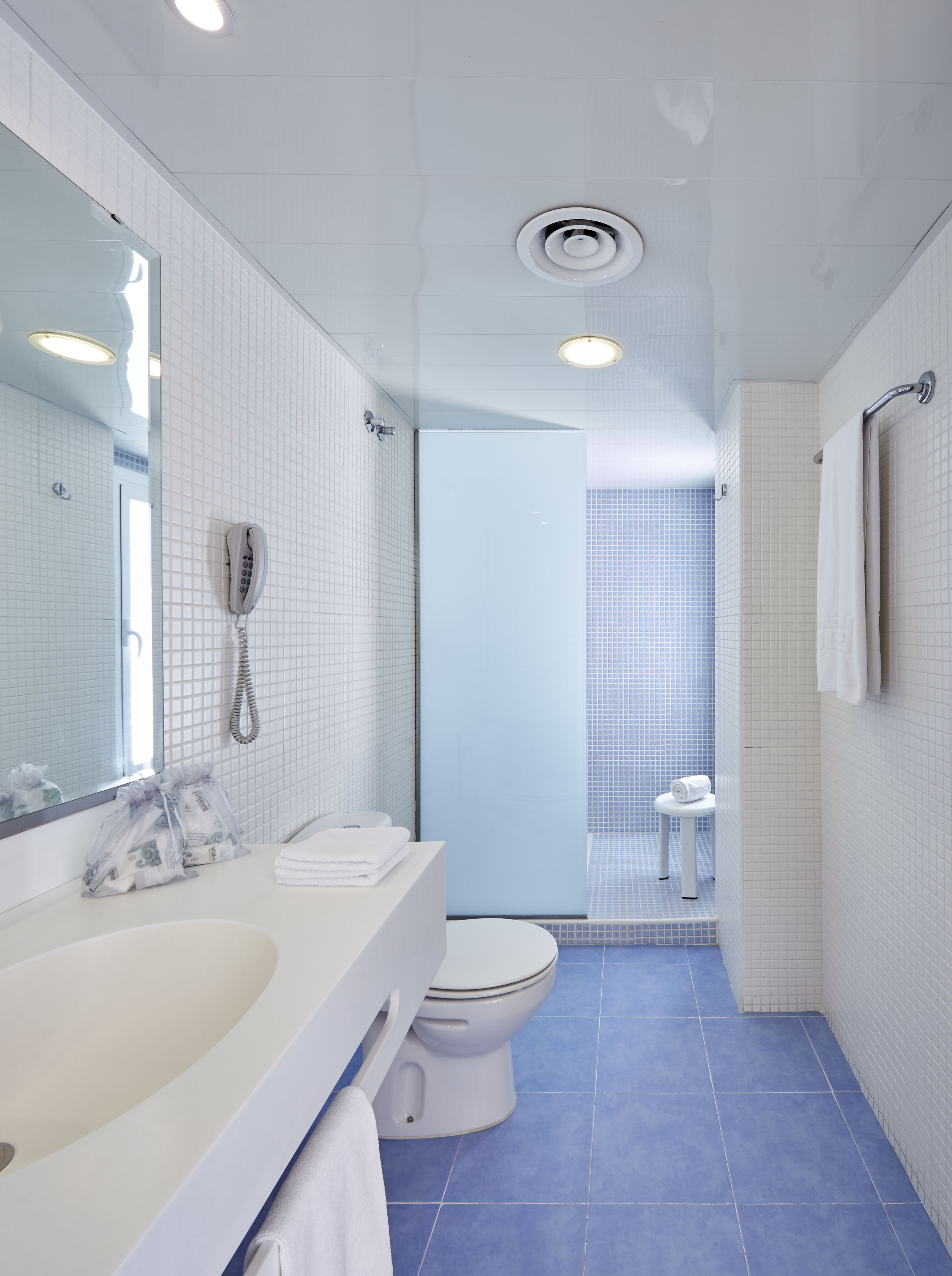 Un baño moderno con lavamanos, inodoro y bidé blancos, un espejo grande y una zona de ducha con azulejos azules.