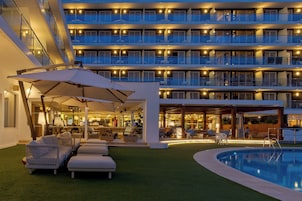 Un hotel de varios pisos con balcones, zona de piscina y mobiliario exterior.