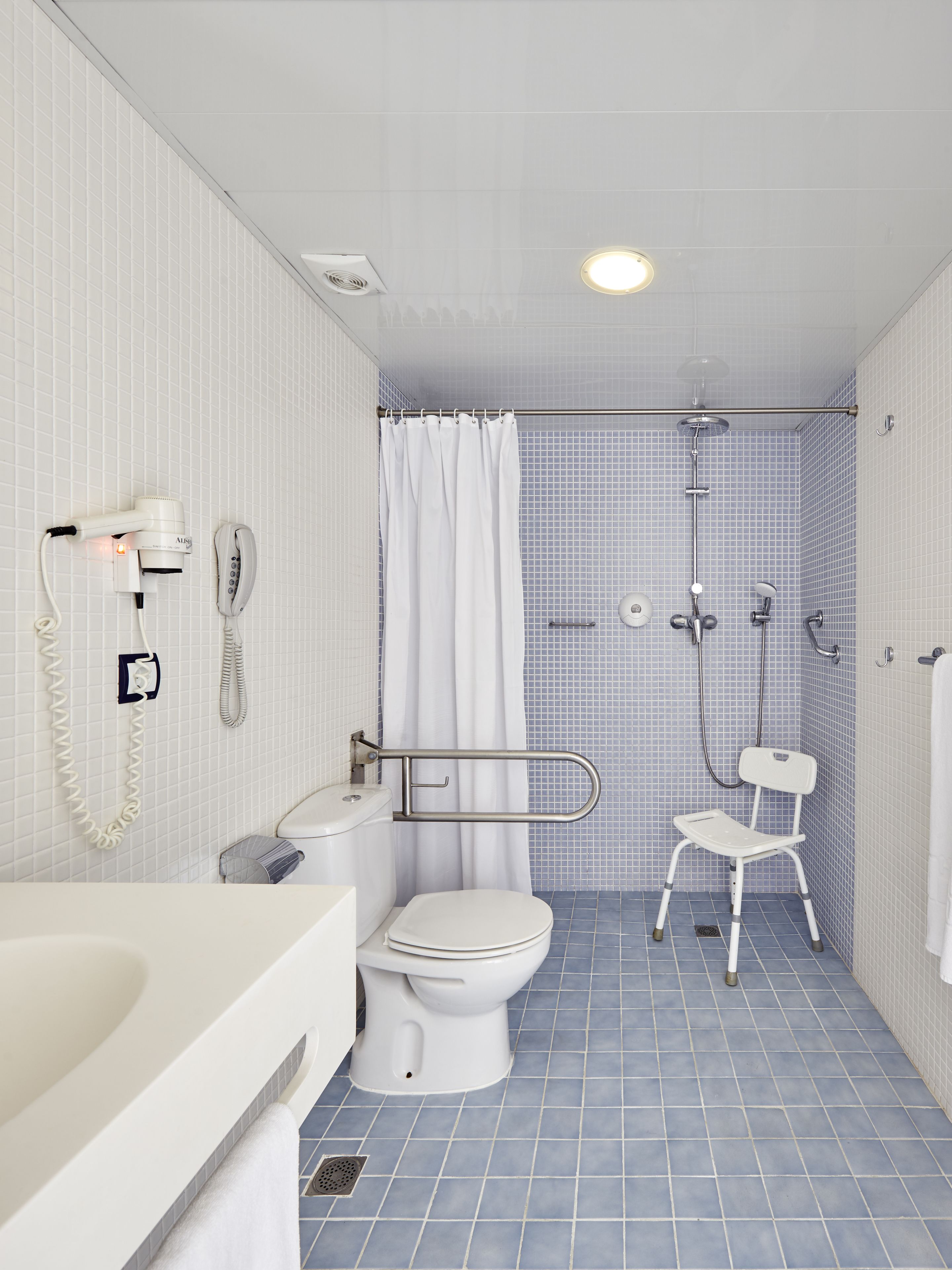 A bathroom with a toilet, a shower with a handheld showerhead, a white chair, and a telephone mounted on the wall.
