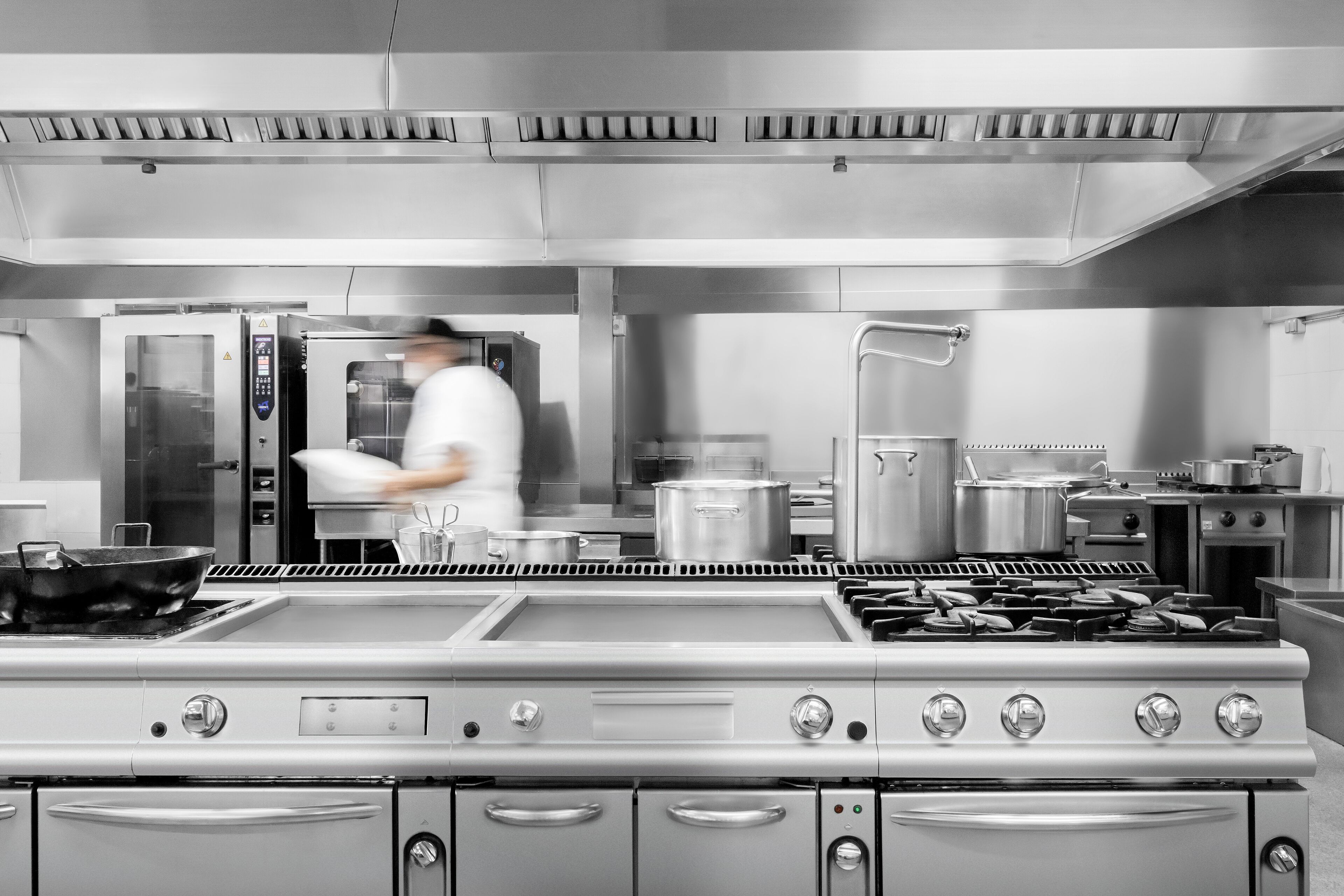 Una cocina comercial con electrodomésticos de acero inoxidable, incluyendo una estufa y un horno grandes, y un chef en movimiento.