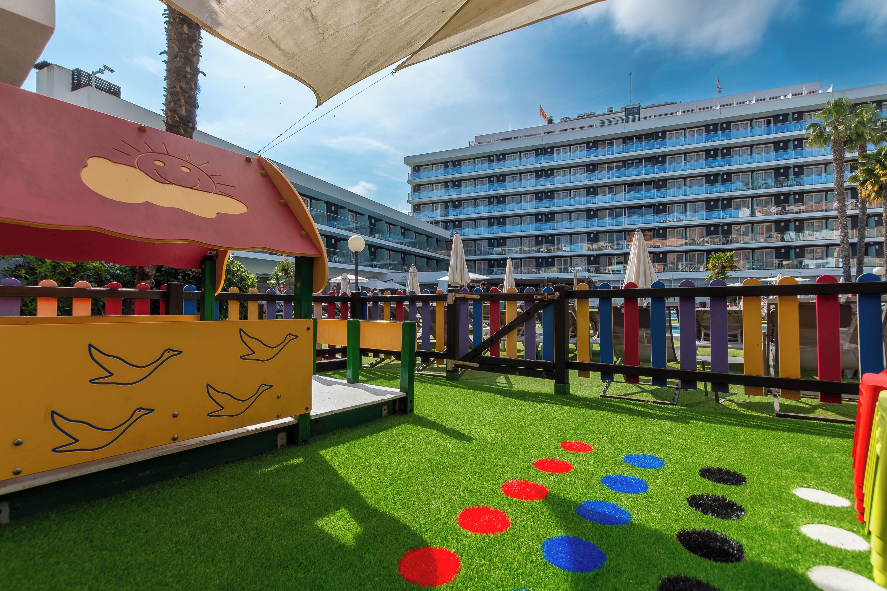 A children's play area with a slide and climbing structure, surrounded by artificial grass and colorful circular patterns.