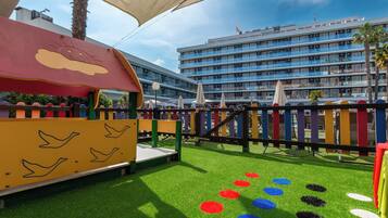 Un espace de jeux pour enfants avec un toboggan et une structure d’escalade, entouré de gazon synthétique et de motifs circulaires colorés.