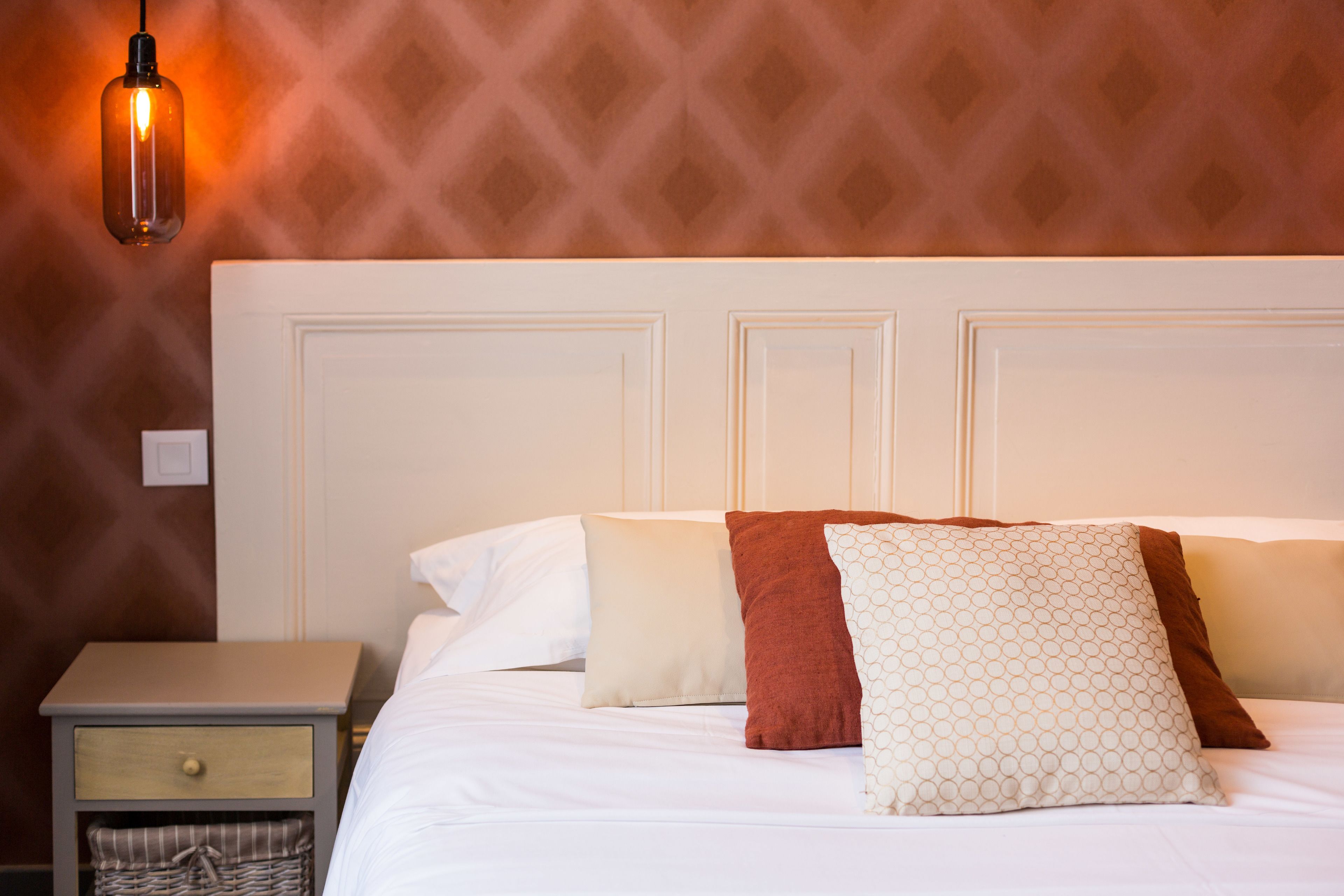 A neatly made bed with white and patterned pillows, a wooden nightstand with a drawer, and a wall-mounted light fixture.