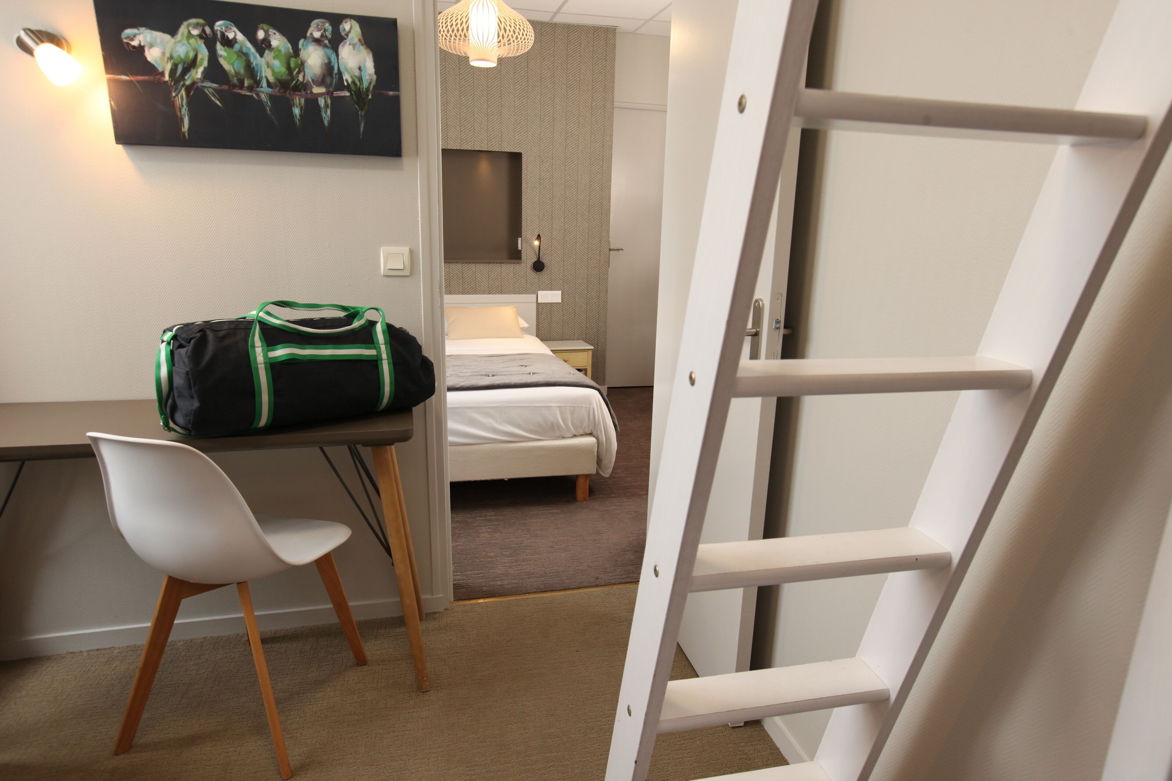 Habitación de hotel pequeña con cama, escritorio con silla, un cuadro en la pared con loros y una escalera.