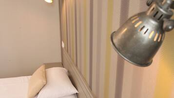 A hotel room with two beds, each with white linens and beige pillows. There is a wall-mounted lamp on the right side of the image.