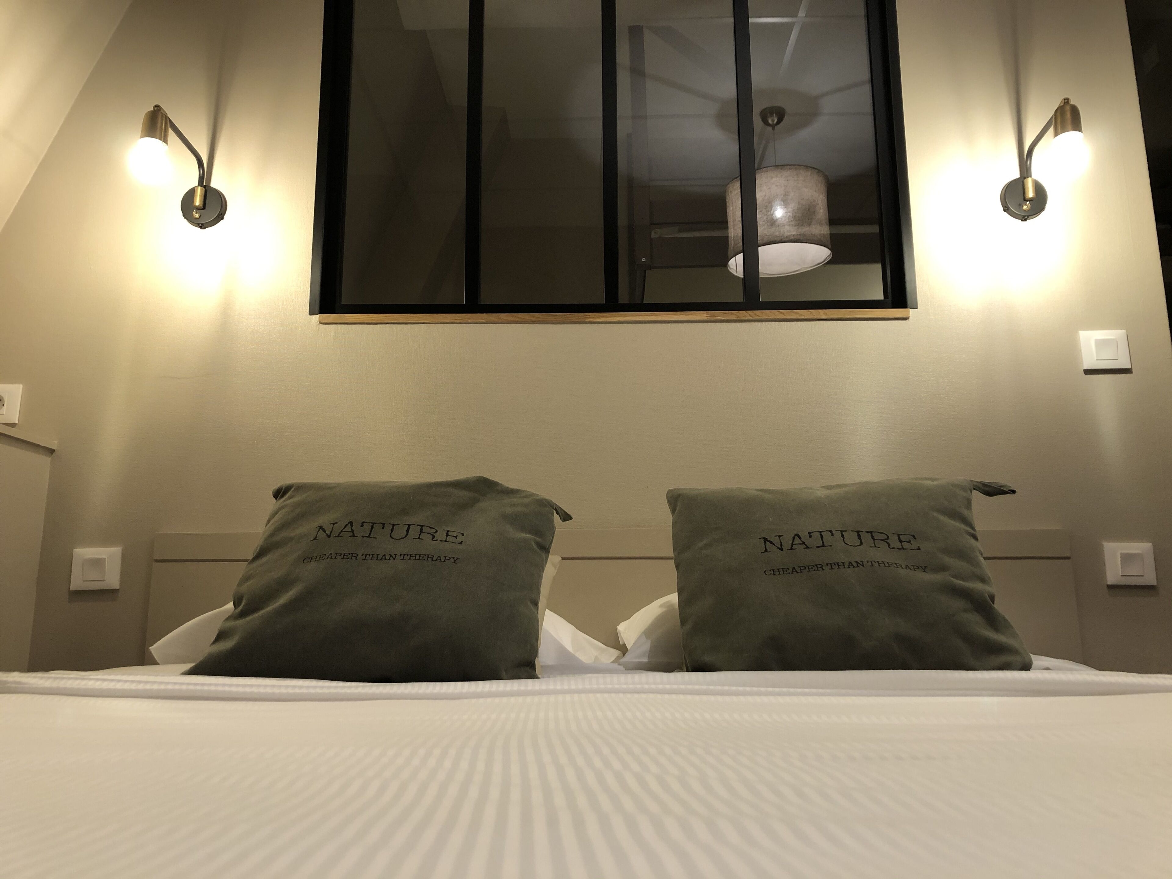 A hotel room with two pillows labeled "NATURE CHEAPER THAN THERAPY" on a bed with white bedding.