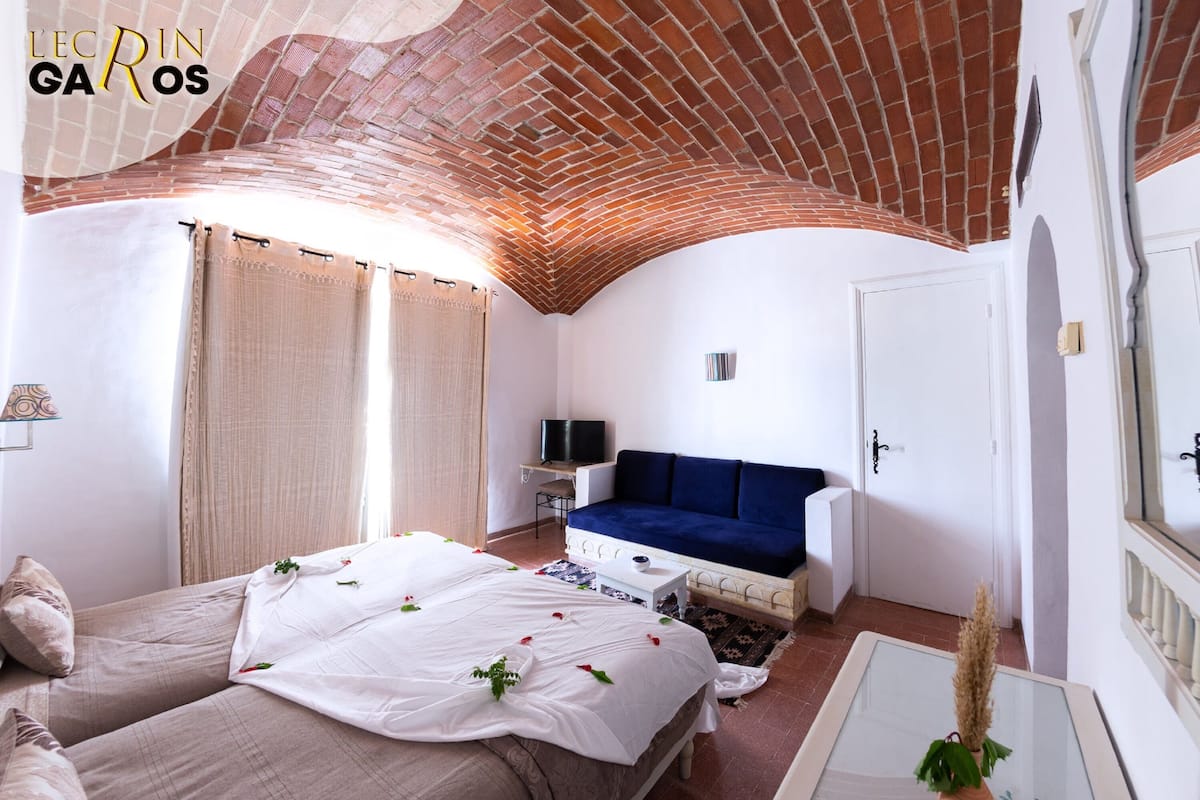 A hotel room with a brick ceiling, a bed with white linens, a blue sofa, and a small table with a plant.