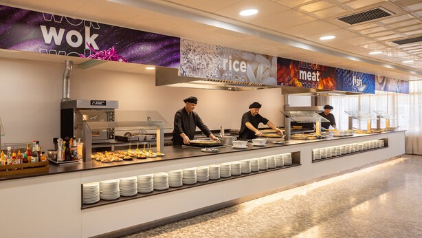 Zona de bufé con un mostrador que exhibe diferentes platos, un cartel grande con categorías de comida y un interior moderno y limpio.