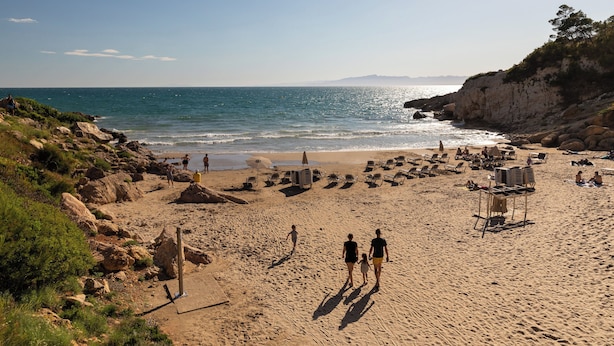 Una playa arenosa con afloramientos rocosos, algunas personas caminando y sillas y sombrillas de playa.