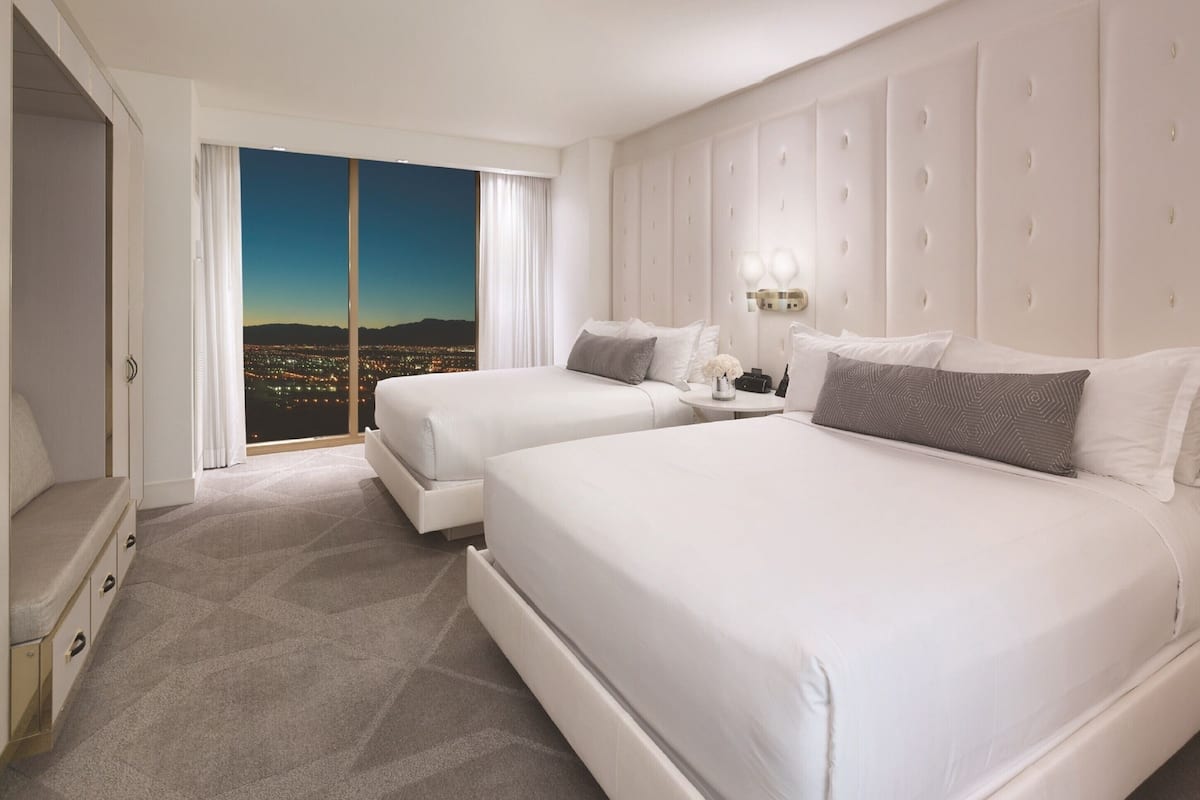 A hotel room with two beds, a large window with a city view, a white headboard, and a bedside table with a lamp.