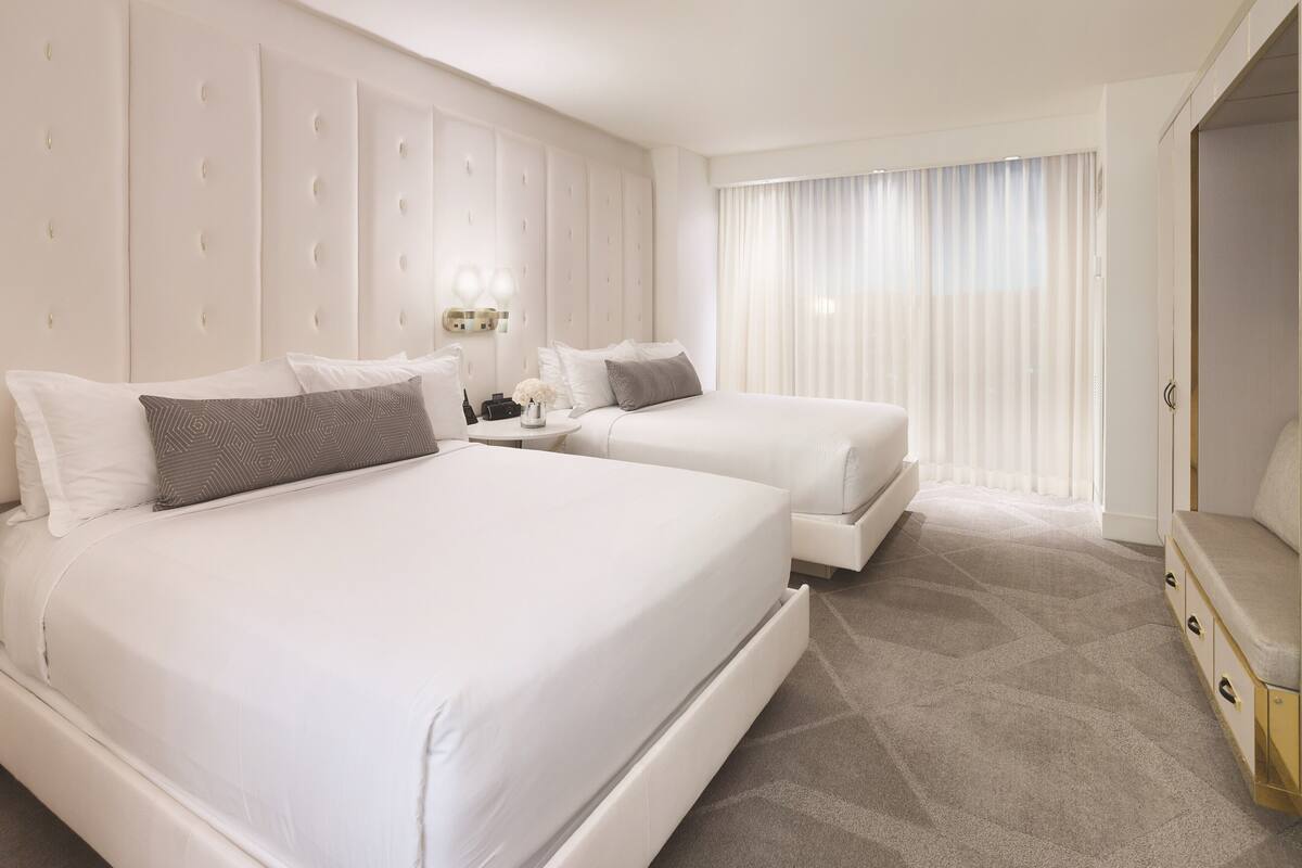 A hotel room with two beds, a large padded headboard, a vanity with a mirror, and a large window with sheer curtains.