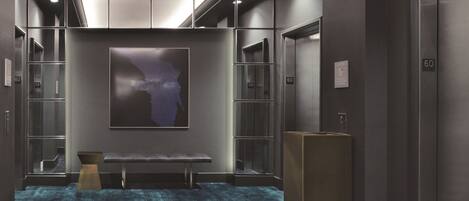 A modern elevator lobby with a blue carpet, a bench, and a large abstract painting on the wall.