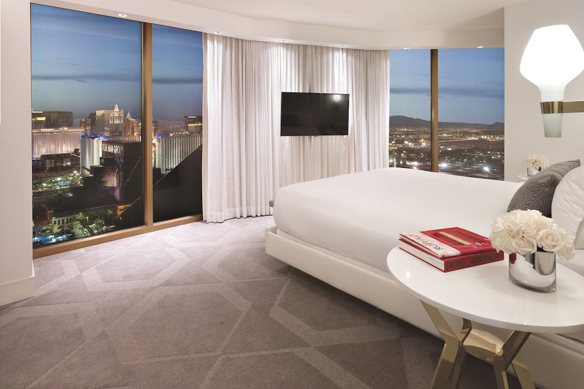 A modern hotel room with a large bed, a flat-screen TV, a round table with a vase of flowers, and a view of a cityscape at night.