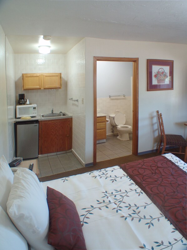 A compact hotel room with a kitchenette, a single bed, a chair, and a bathroom visible through an open door.