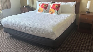 A neatly made bed with a white bedspread, two pillows with a floral design, a wooden nightstand with a lamp, and a carpeted floor.