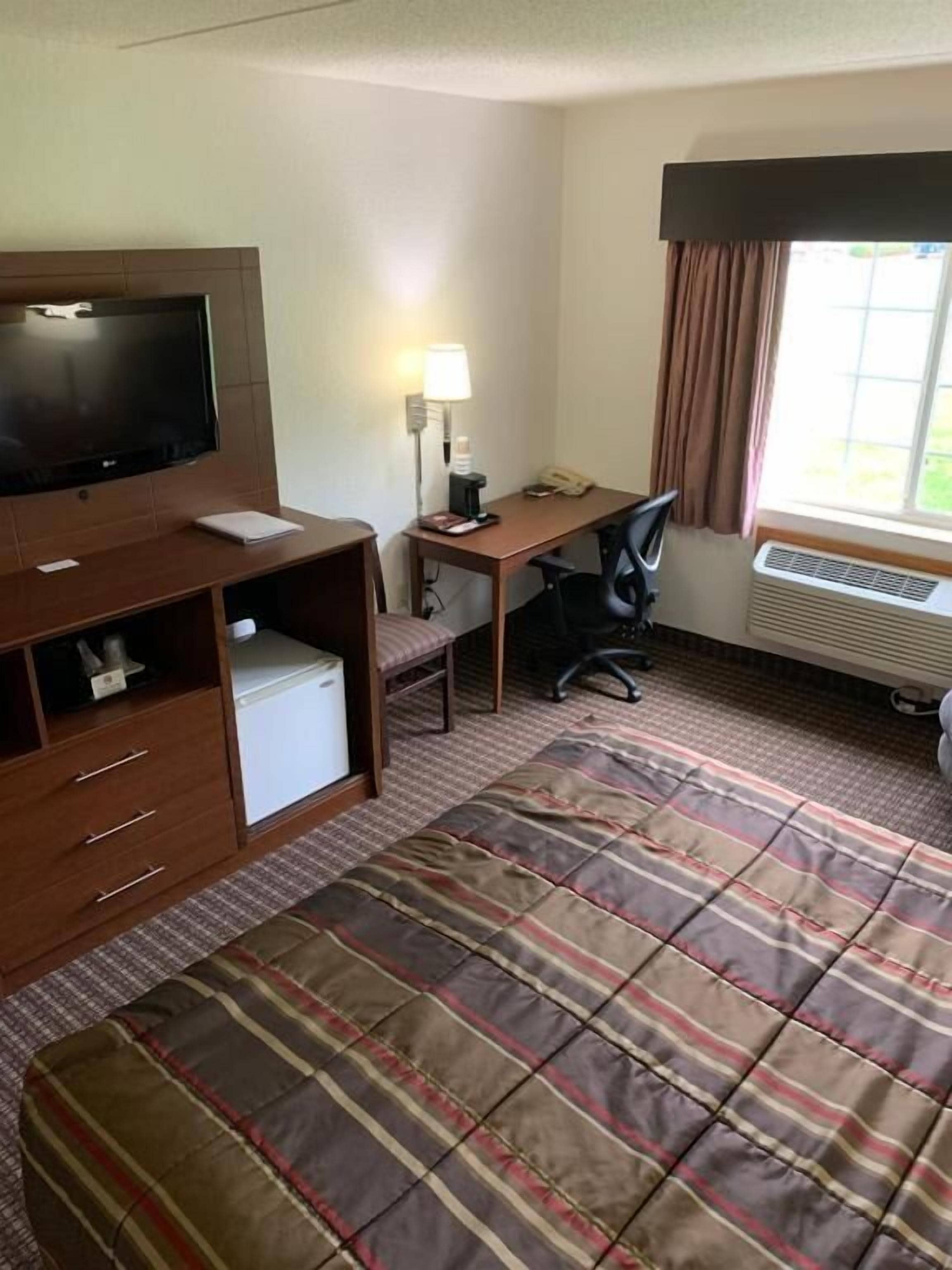 Habitación de hotel con cama, escritorio, silla, televisor, lámpara y ventana con cortinas.