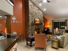 Un vestíbulo de hotel con chimenea de piedra, una recepción, un área de descanso con sofá y sillones, y un amplio ventanal con cortinas.