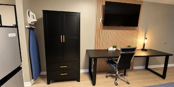 Una habitación con un armario negro, un escritorio, una silla, un televisor de pantalla plana montado en un panel de madera y una mesita de noche con una lámpara.