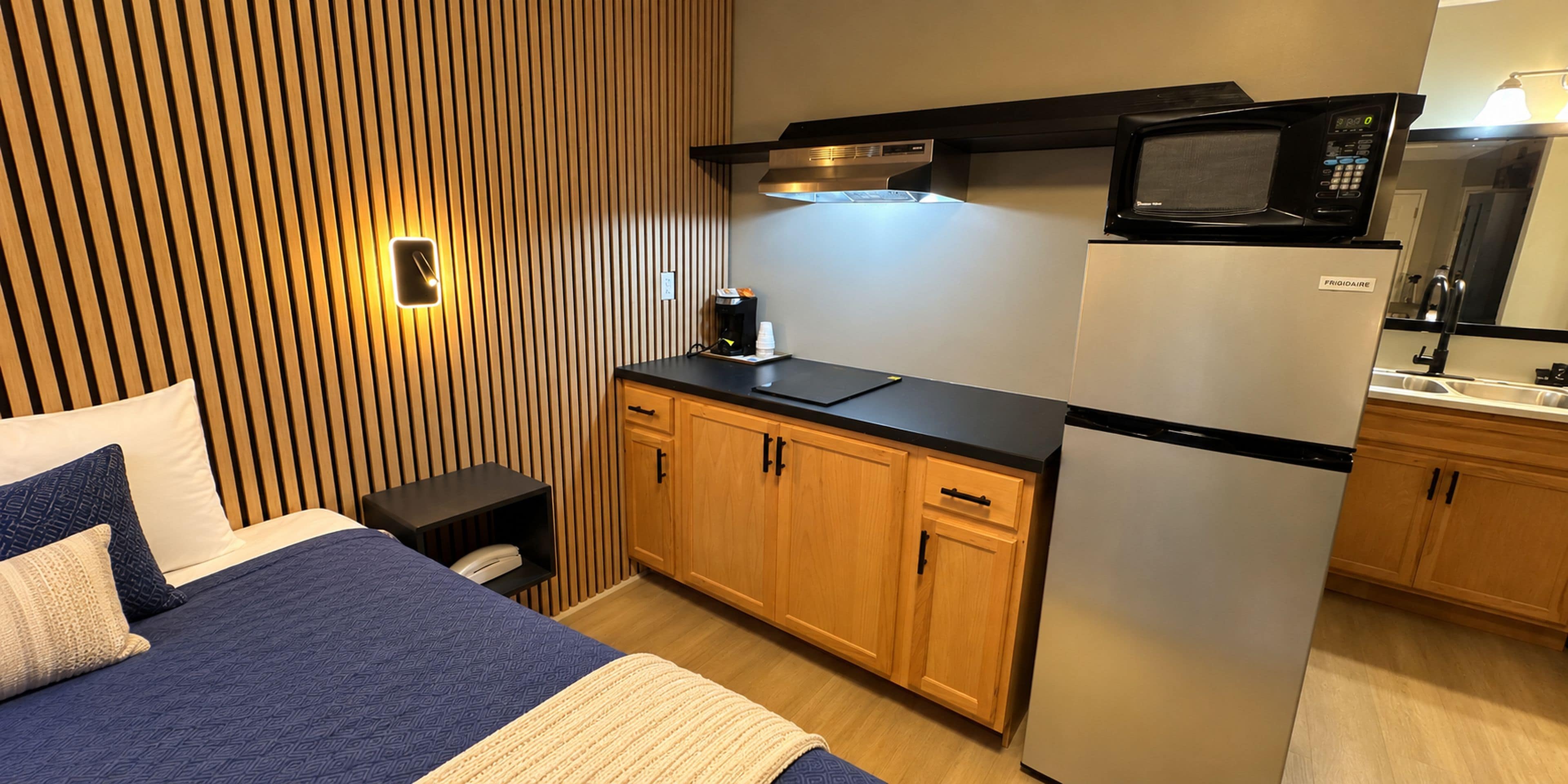 A compact hotel room with a kitchenette, a bed with blue bedding, and a wooden slatted wall.