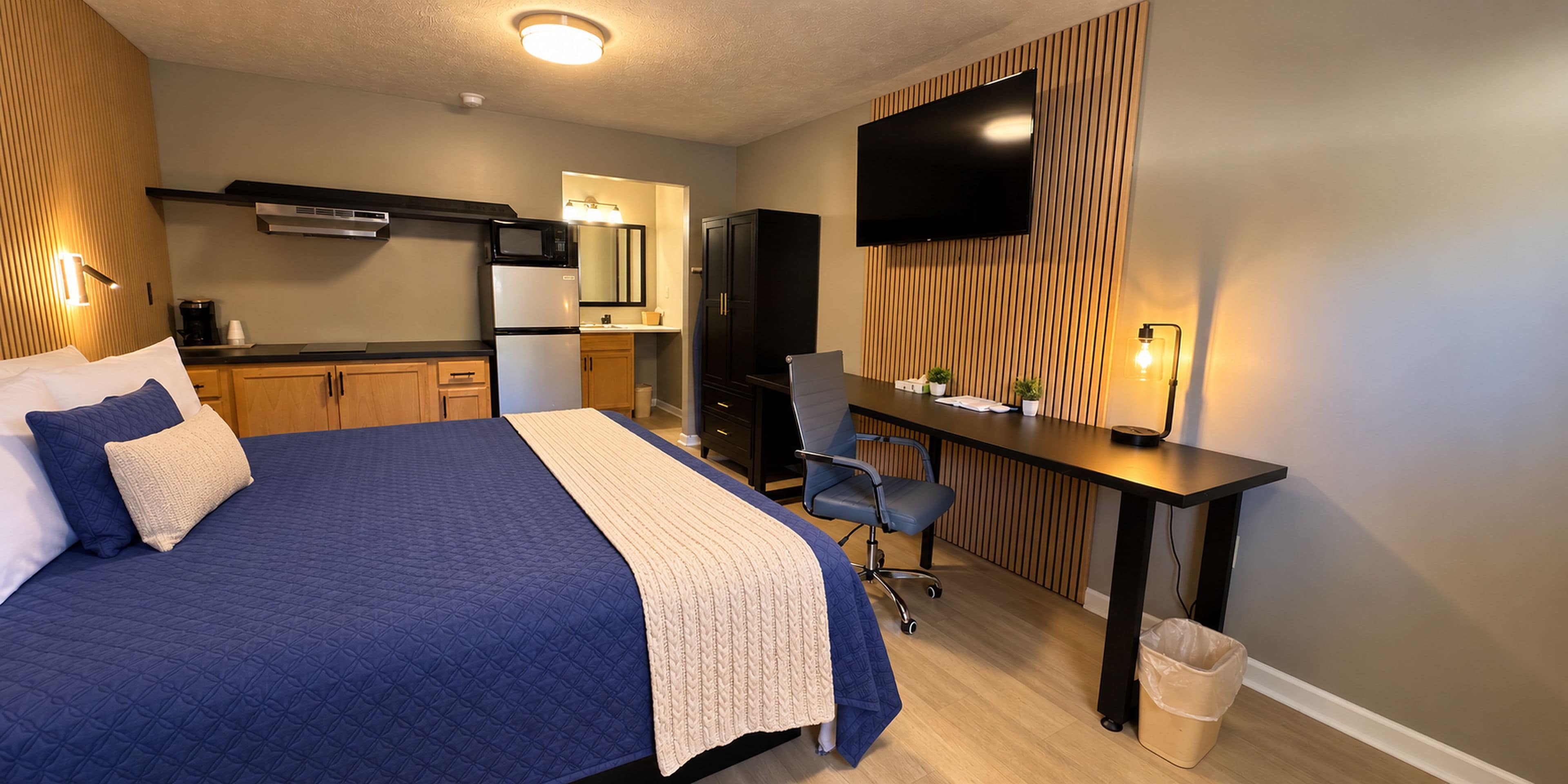 Habitación de hotel con cama, escritorio, televisión, kitchenette y cubo de basura.
