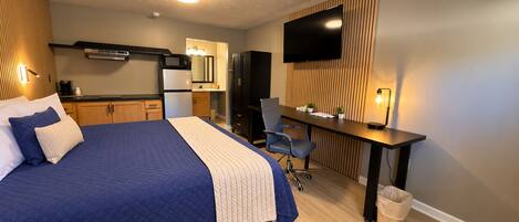 A hotel room with a bed, a desk, a television, a kitchenette, and a waste bin.