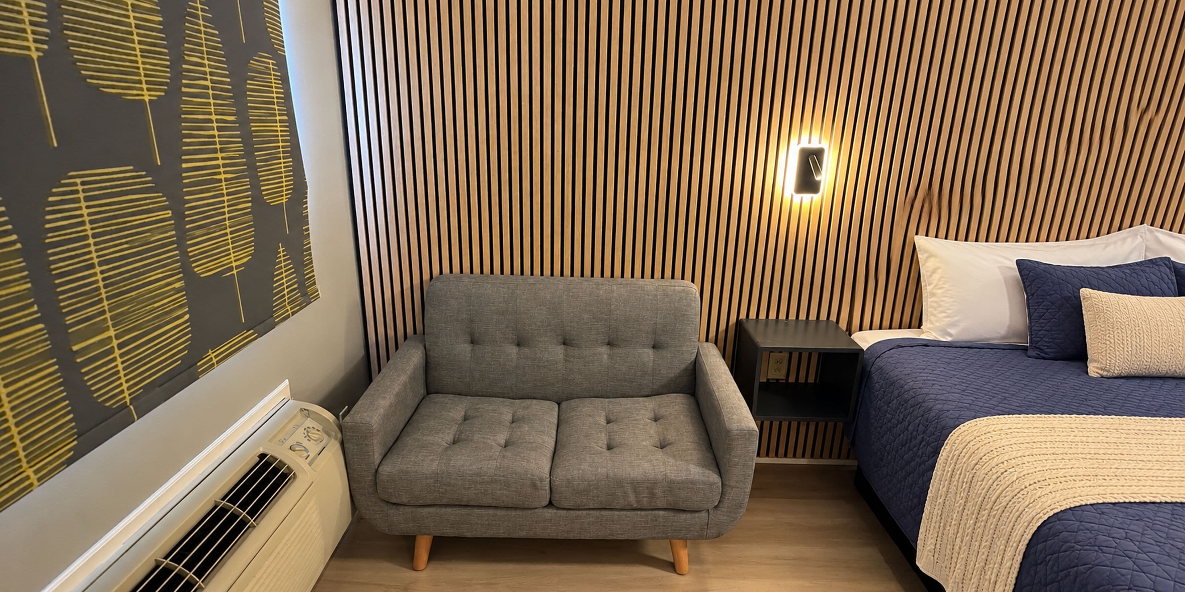 Una habitación con una silla tapizada de gris, una cama con colcha azul oscuro y una pared con listones de madera verticales.