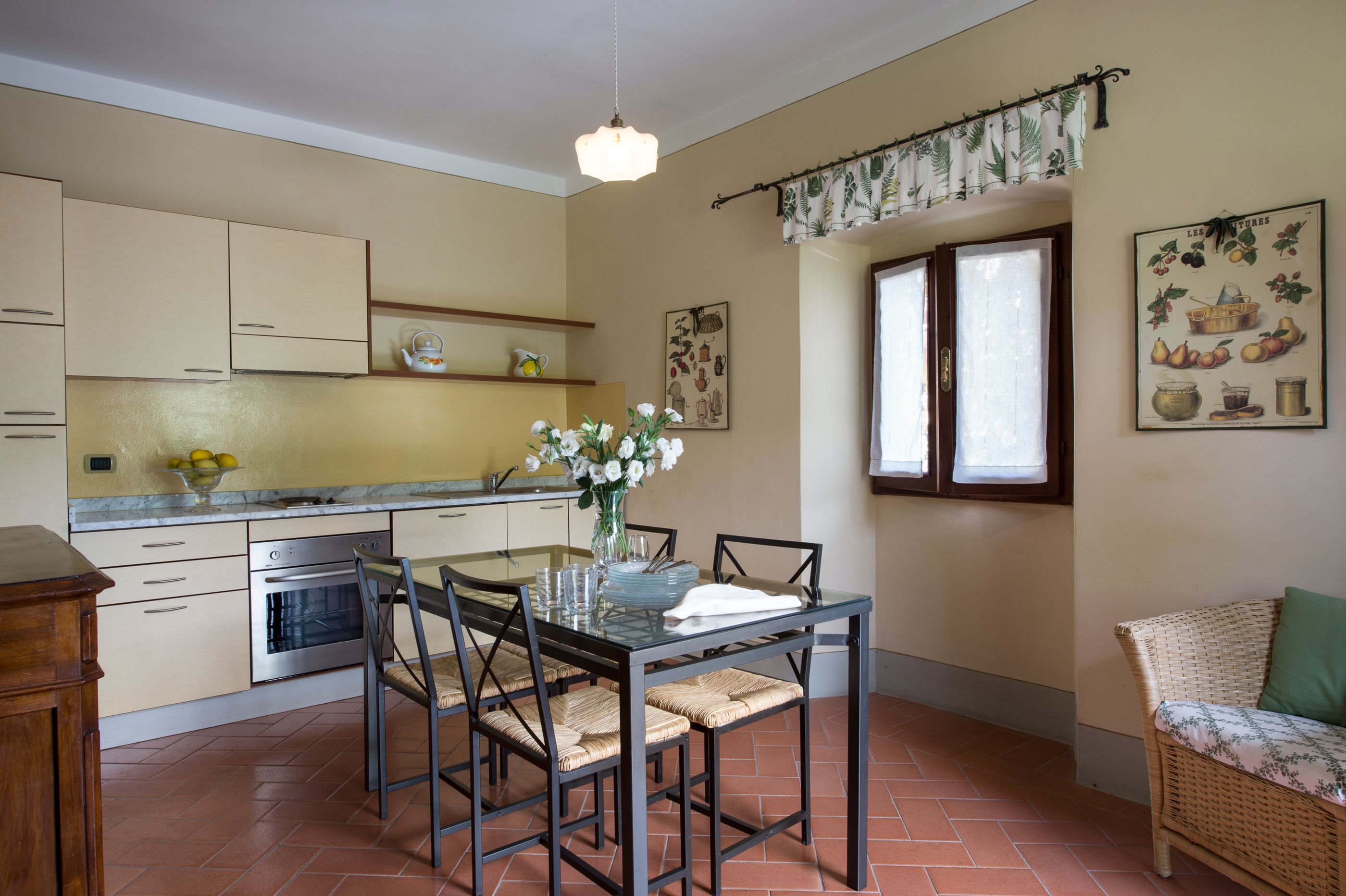 Une cuisine avec une table à manger, des chaises, un vase de fleurs et un fauteuil en osier.
