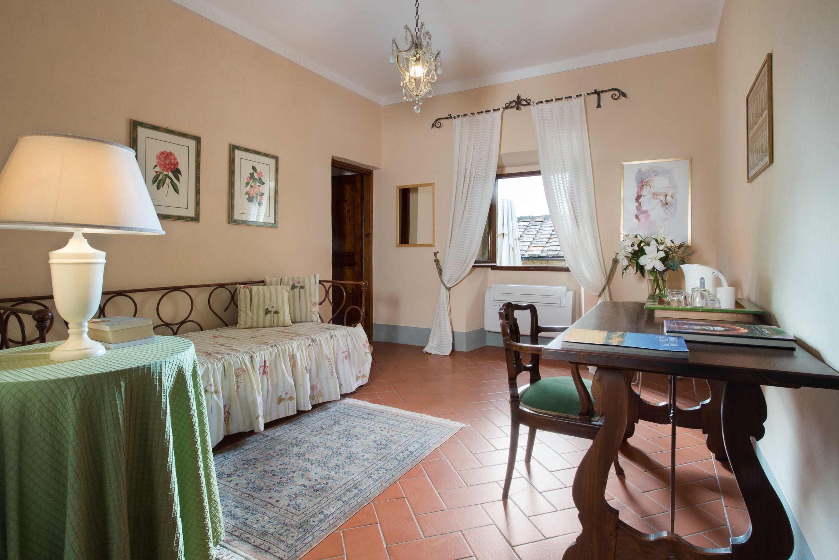 Una habitación con cama, una mesa con lámpara, una silla, una ventana con cortinas y un candelabro.