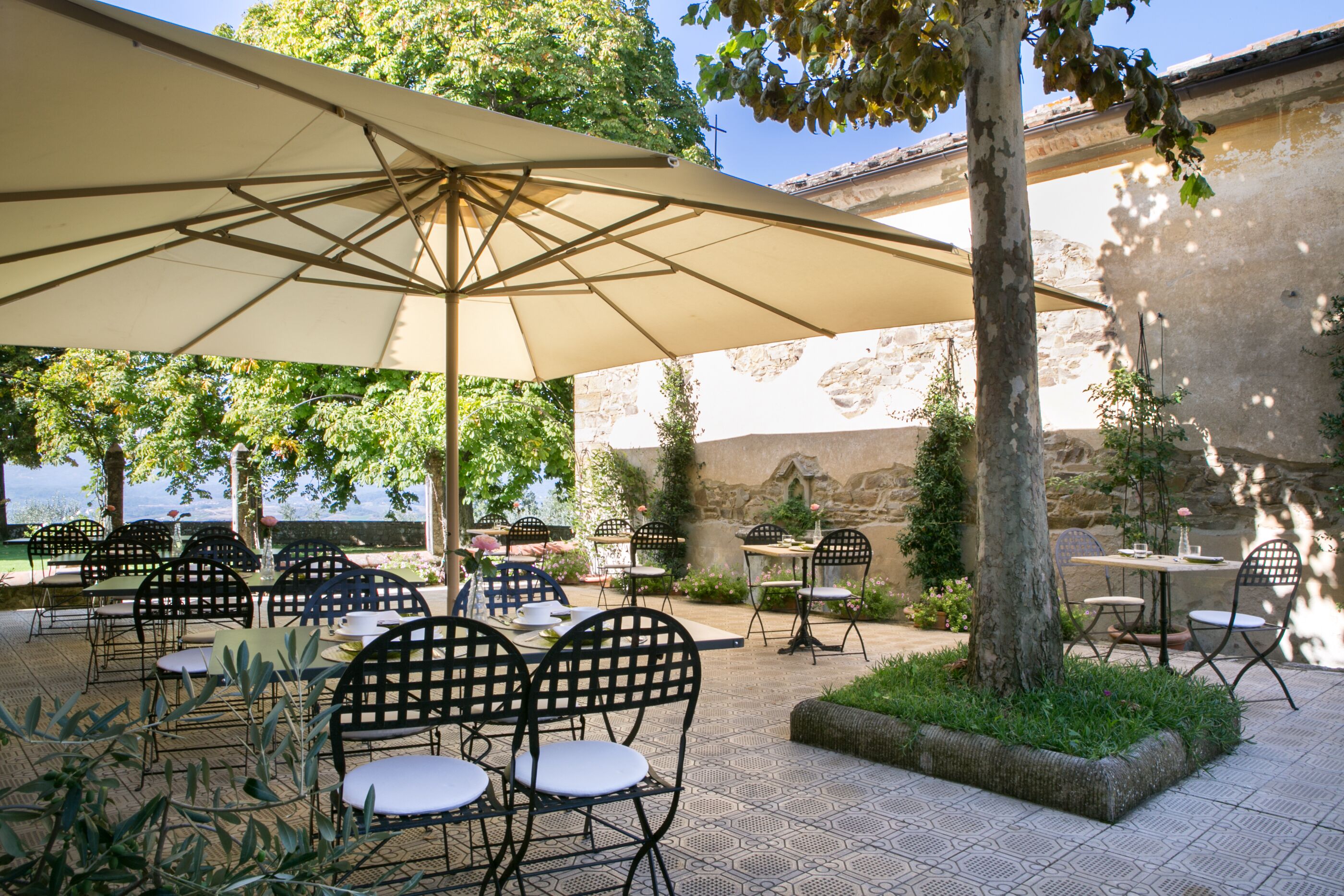 An outdoor patio with tables and chairs, a large umbrella, and a view of the sea.