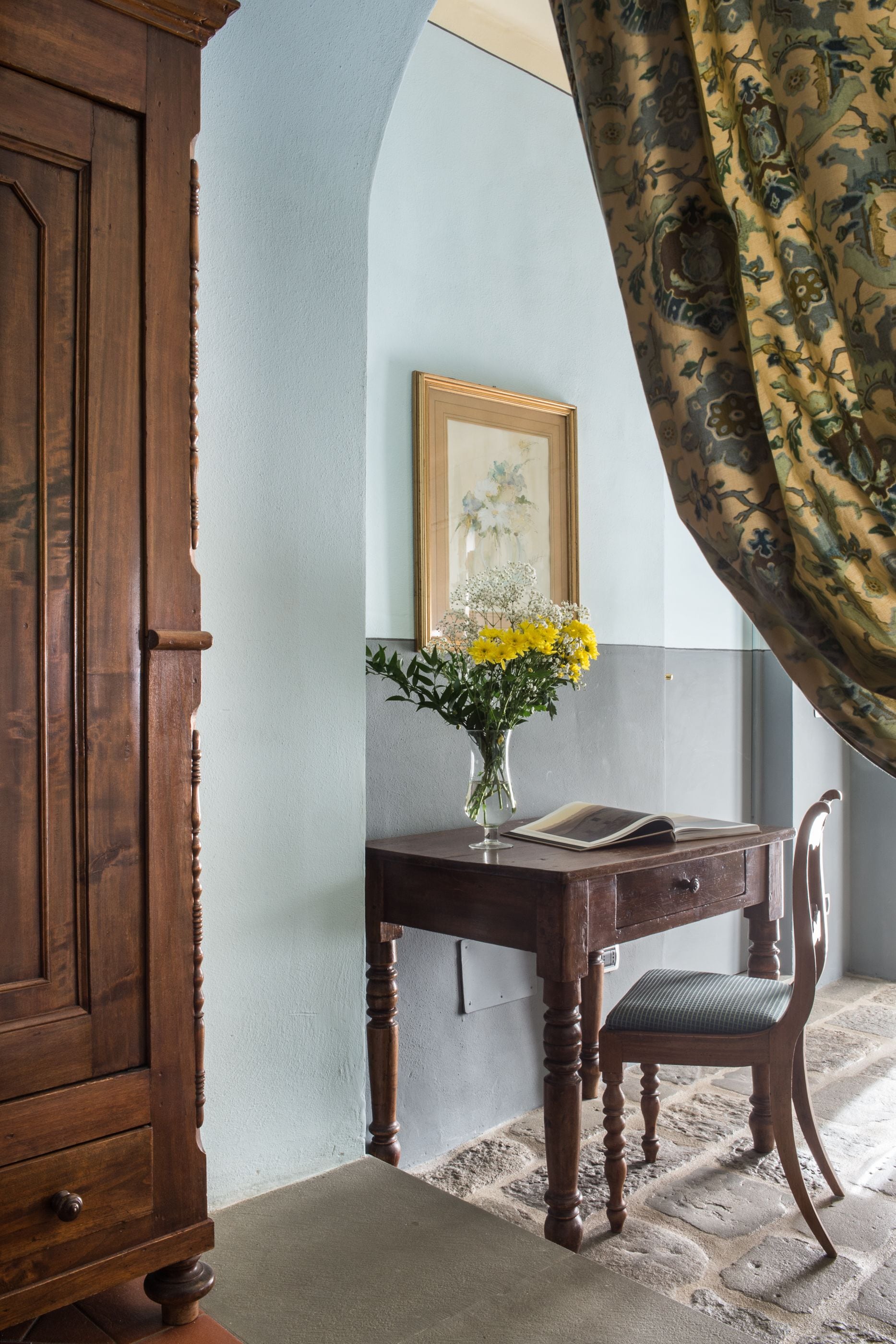 A wooden desk with a vase of yellow flowers, a framed picture on the wall, and a chair.