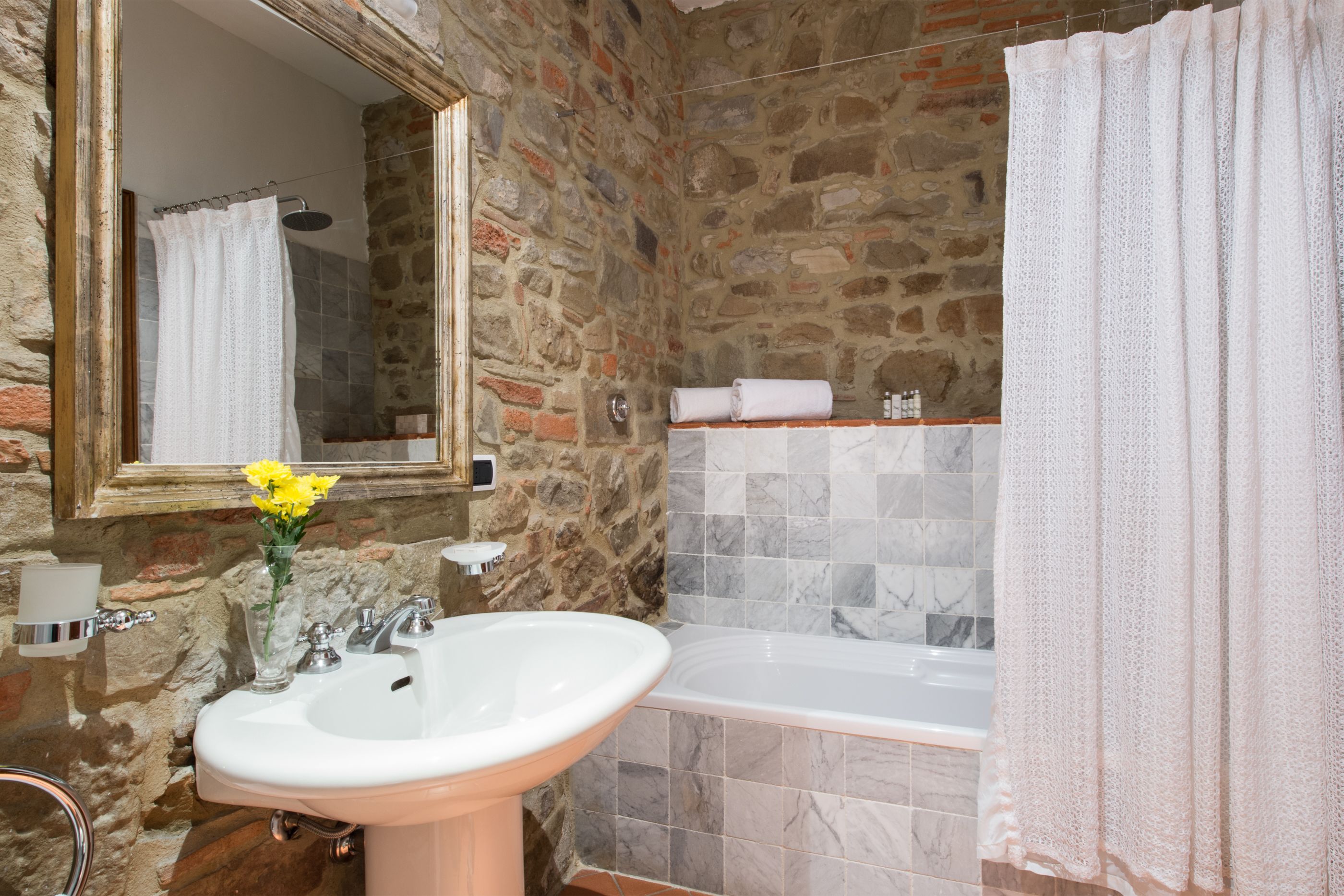 Un baño con pared de piedra, un lavamanos blanco, un espejo, una bañera y una cortina de ducha.
