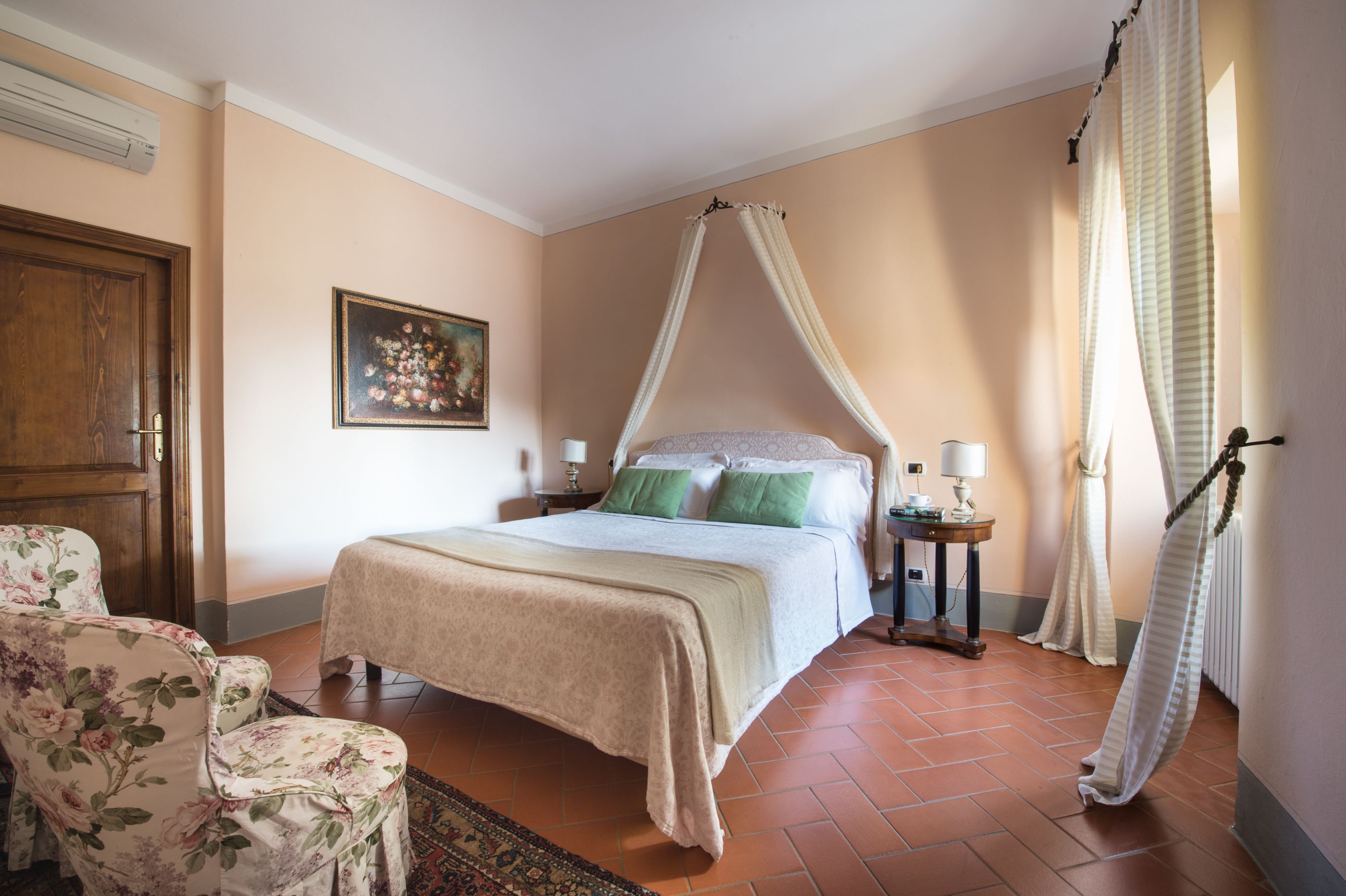 A bedroom with a four-poster bed, a wooden door, a painting on the wall, and a chair with floral upholstery.