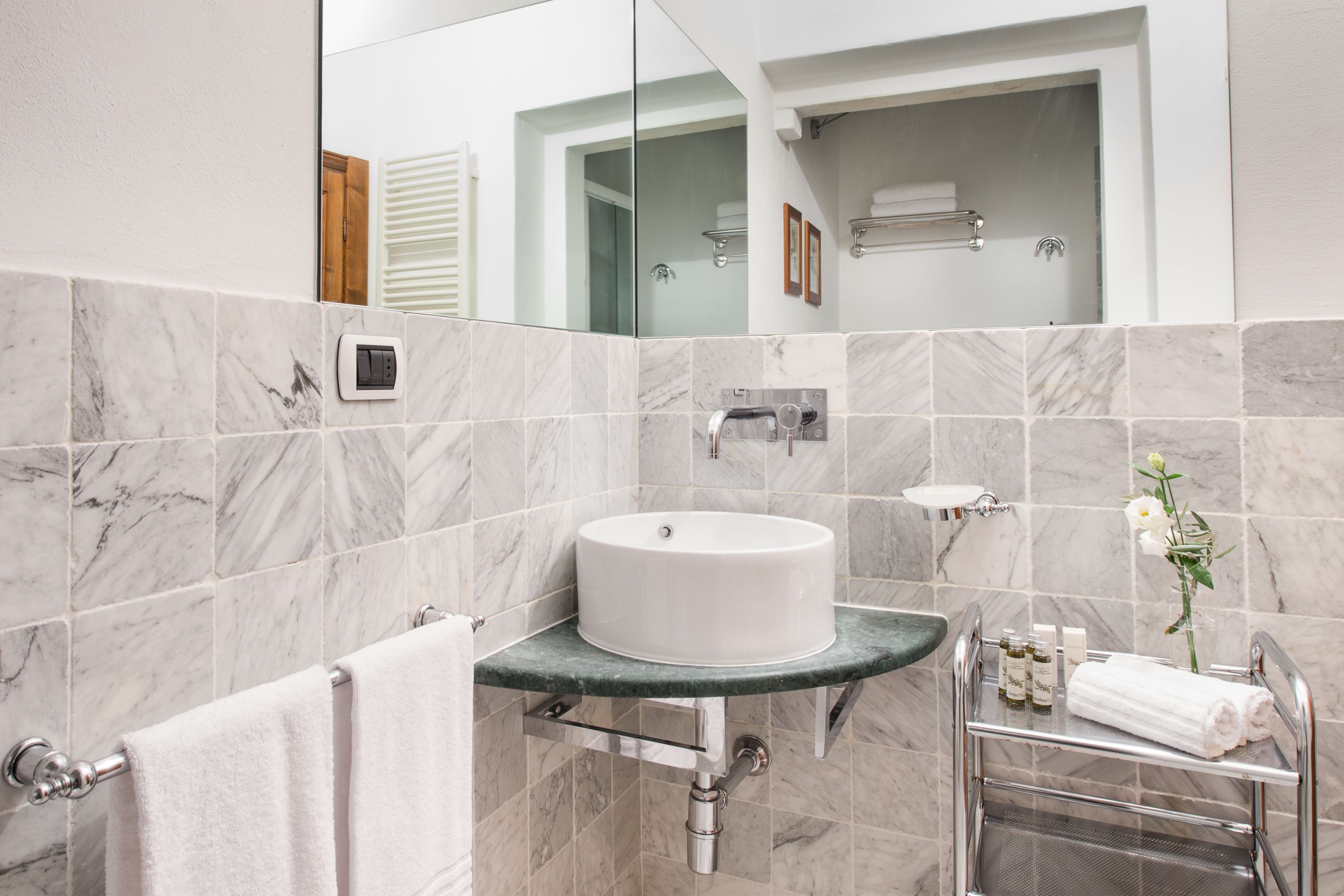 A modern bathroom with a marble-tiled wall, a round sink, a towel rack, and a glass shower enclosure.