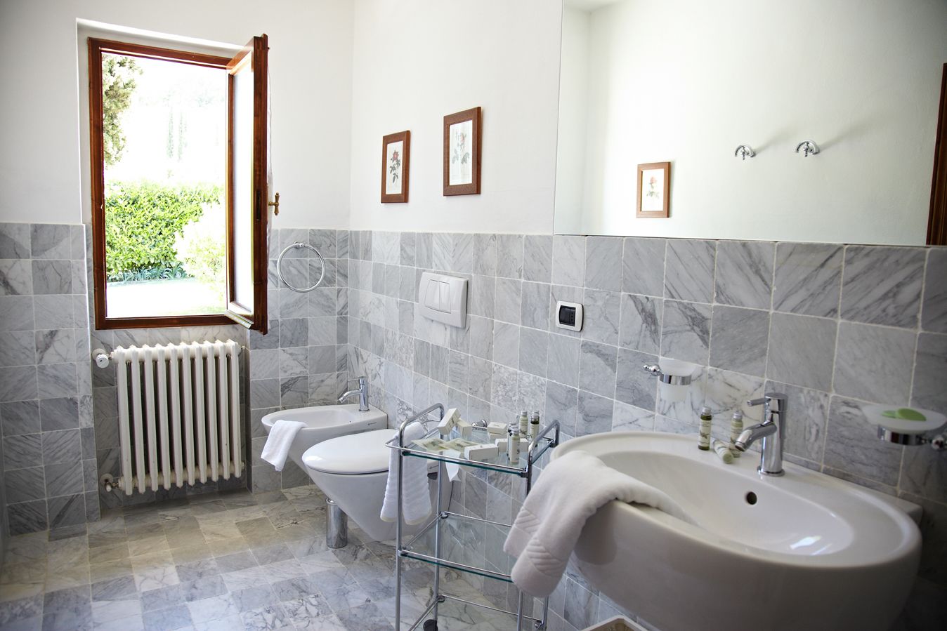 Un baño con dos lavabos, un inodoro, un radiador y una ventana con vistas a zonas verdes.