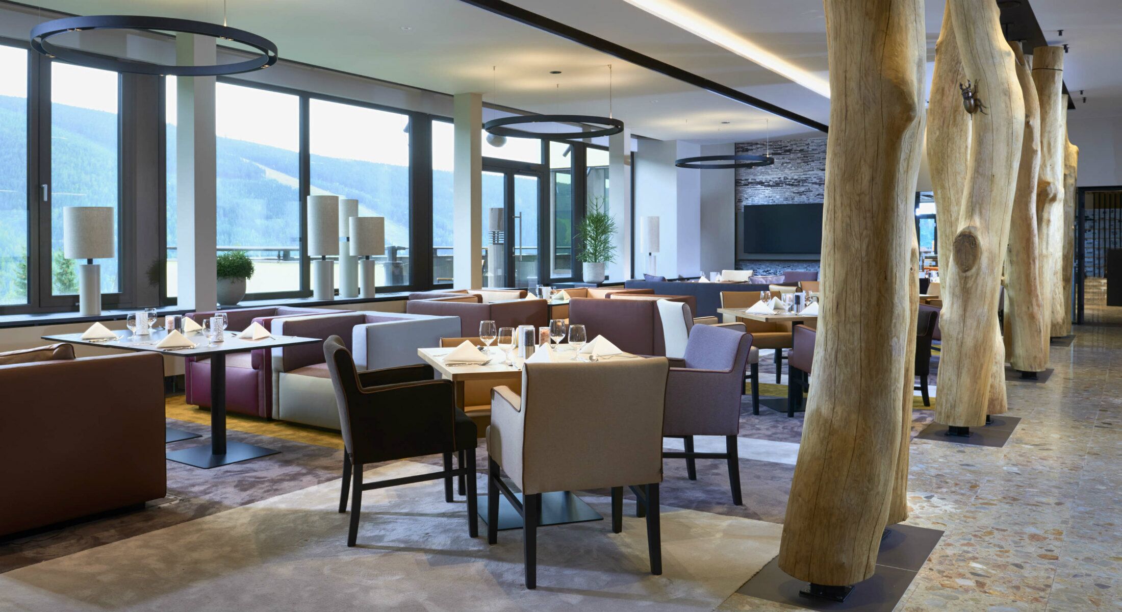 A modern restaurant with high ceilings, large windows, and a view of the mountains. Tables are set with white tablecloths and candles.