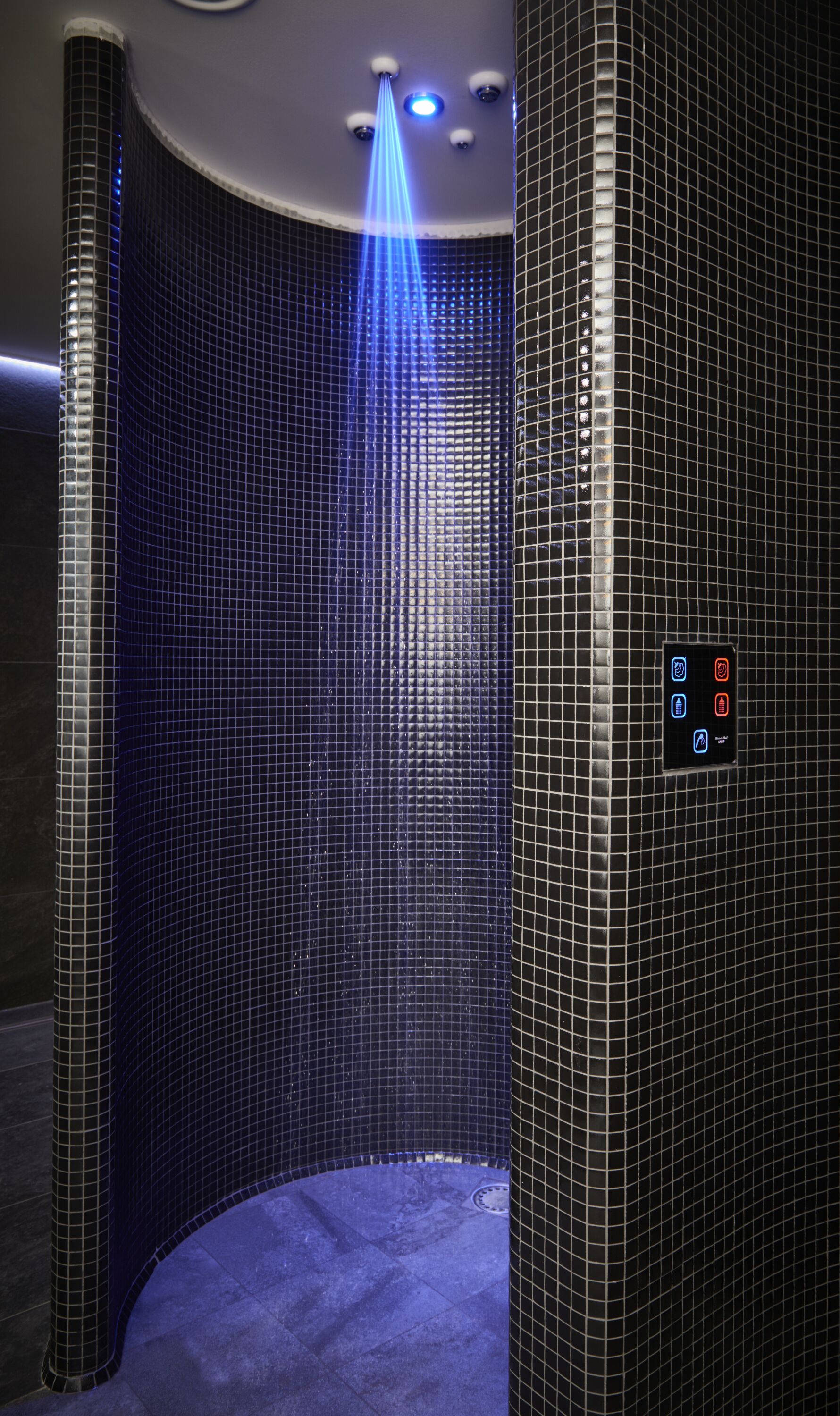 A modern bathroom with a curved design, featuring a blue-tiled wall and a control panel with four illuminated buttons.