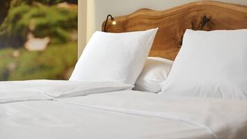 Cama bem arrumada com lençóis brancos e cabeceira de madeira, em frente a um fundo natural desfocado.