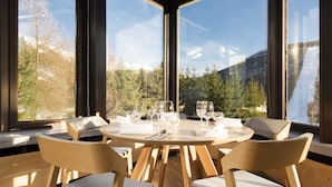 Comedor con una mesa redonda dispuesta para cuatro personas, rodeada de sillas de madera con cojines grises, y ventanas amplias que ofrecen vistas a árboles y montañas.